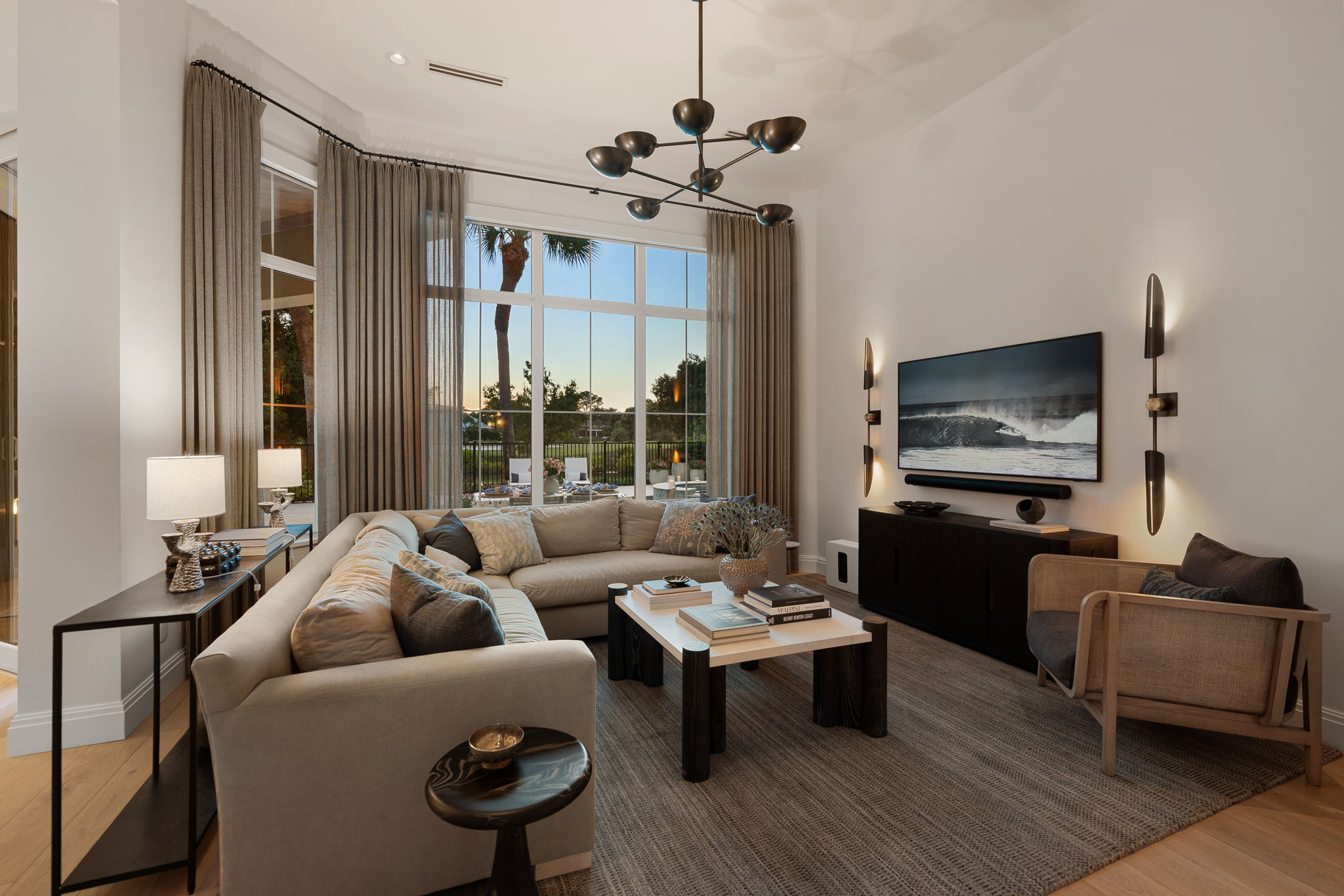 Modern living room with a large beige L-shaped sofa, a wooden armchair with dark cushions, a rectangular coffee table with books and a vase, a dark media console with a wall-mounted TV displaying a wave, two vertical wall sconces, large windows with beige curtains showing an outdoor patio and palm trees at sunset.