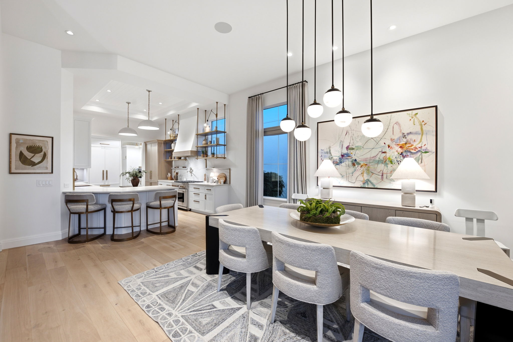Modern open-plan kitchen and dining area with light wood flooring, a large beige dining table surrounded by eight gray upholstered chairs, a decorative rug under the table, and a centerpiece plant. The kitchen features white cabinetry, a large island with three bar stools, stainless steel appliances, open shelving with decorative items, and large windows with gray curtains. Several pendant lights hang above the dining table, and two table lamps illuminate a sideboard beneath a large abstract colorful painting.