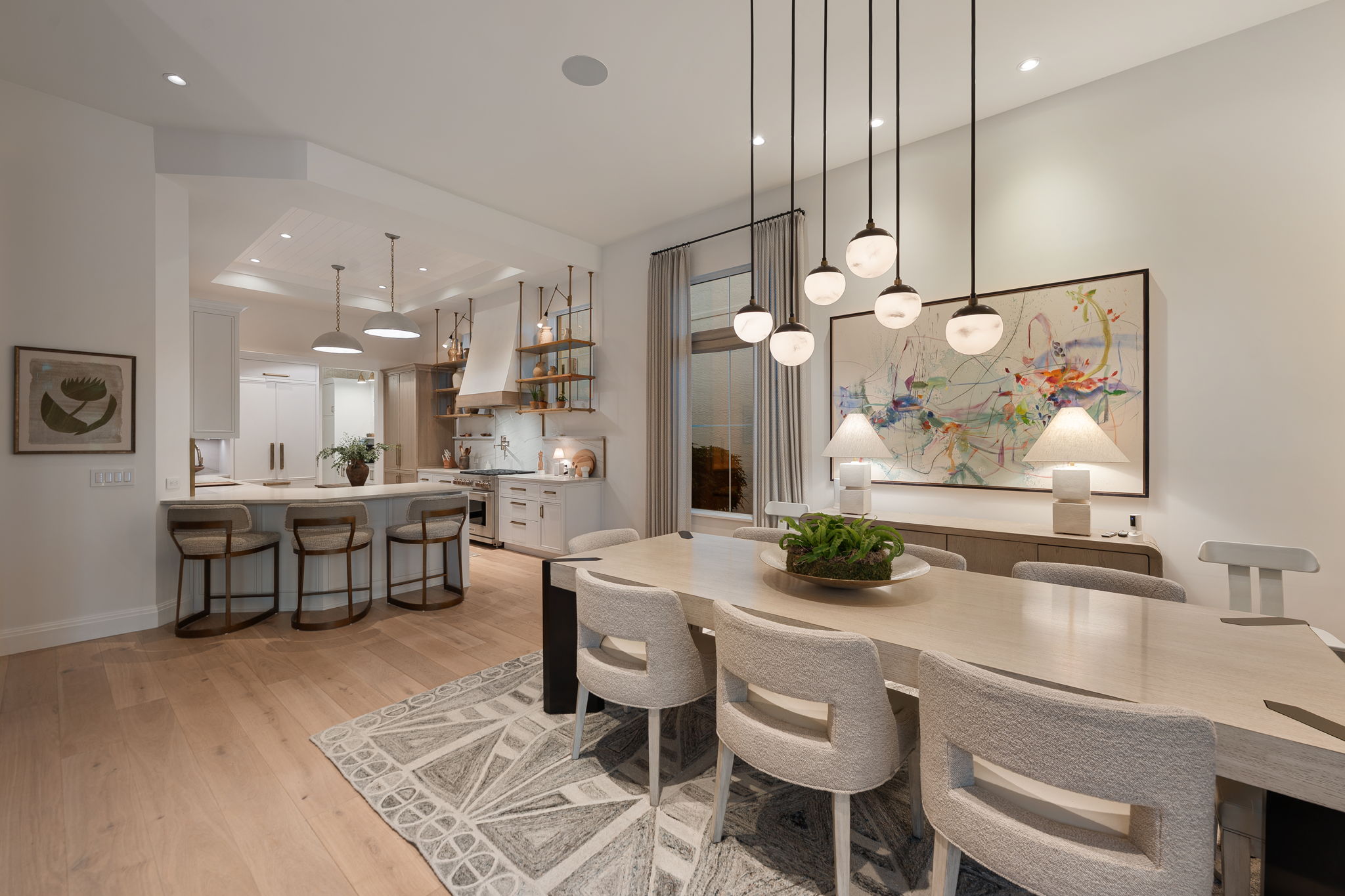 Modern dining area and kitchen with light wood flooring, beige upholstered chairs around a light wood dining table on a patterned rug, pendant lights hanging above, a large abstract painting on the wall, two table lamps on a sideboard, and a kitchen with white cabinetry, open shelves, and three counter stools at an island.