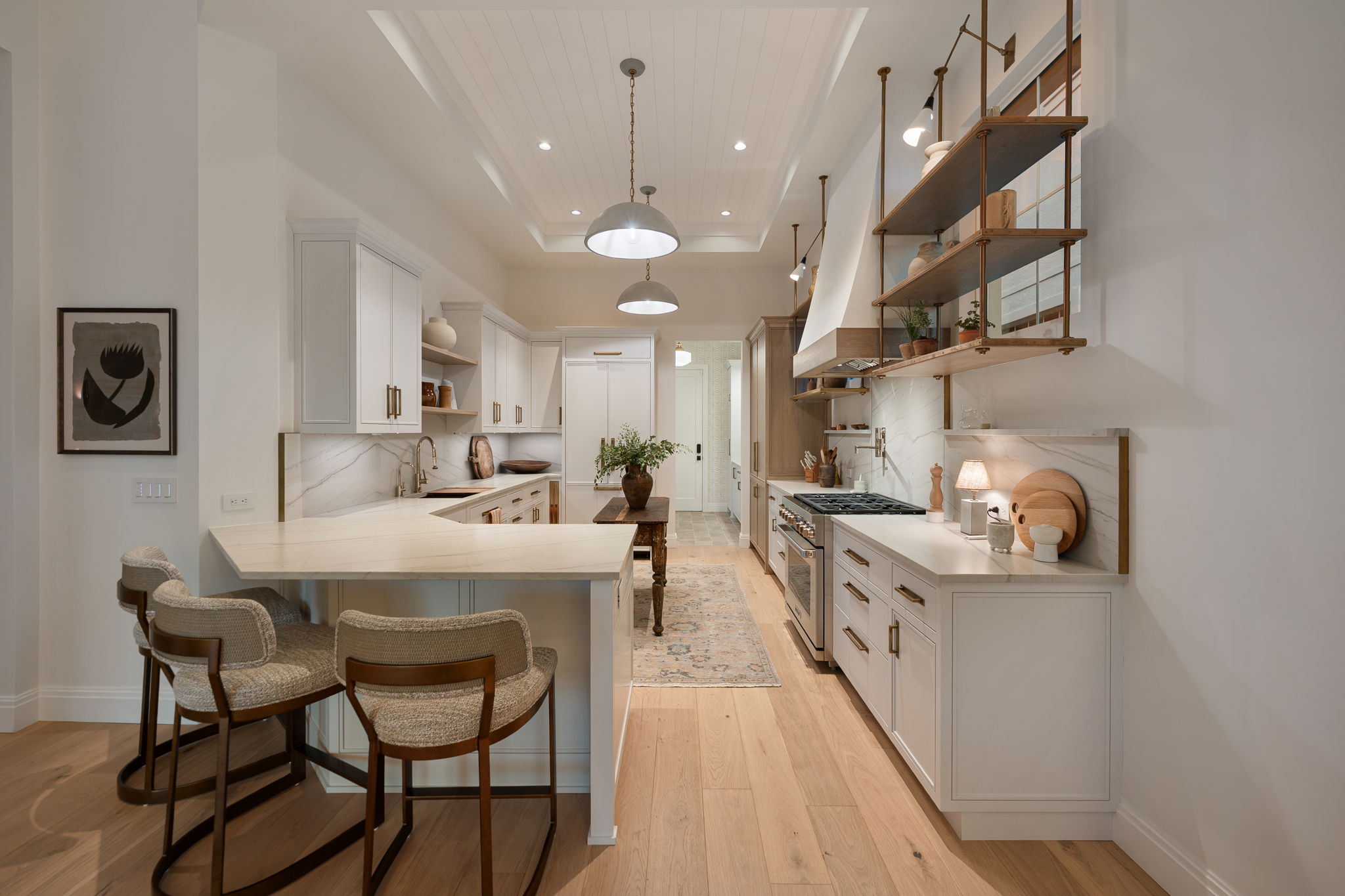 Modern kitchen with white cabinetry, marble countertops and backsplash, wooden floor, and a wooden island table with a vase of greenery in the center; three cushioned bar stools at a peninsula counter; hanging metal pendant lights and open wooden shelves with plants and decor on the right wall.