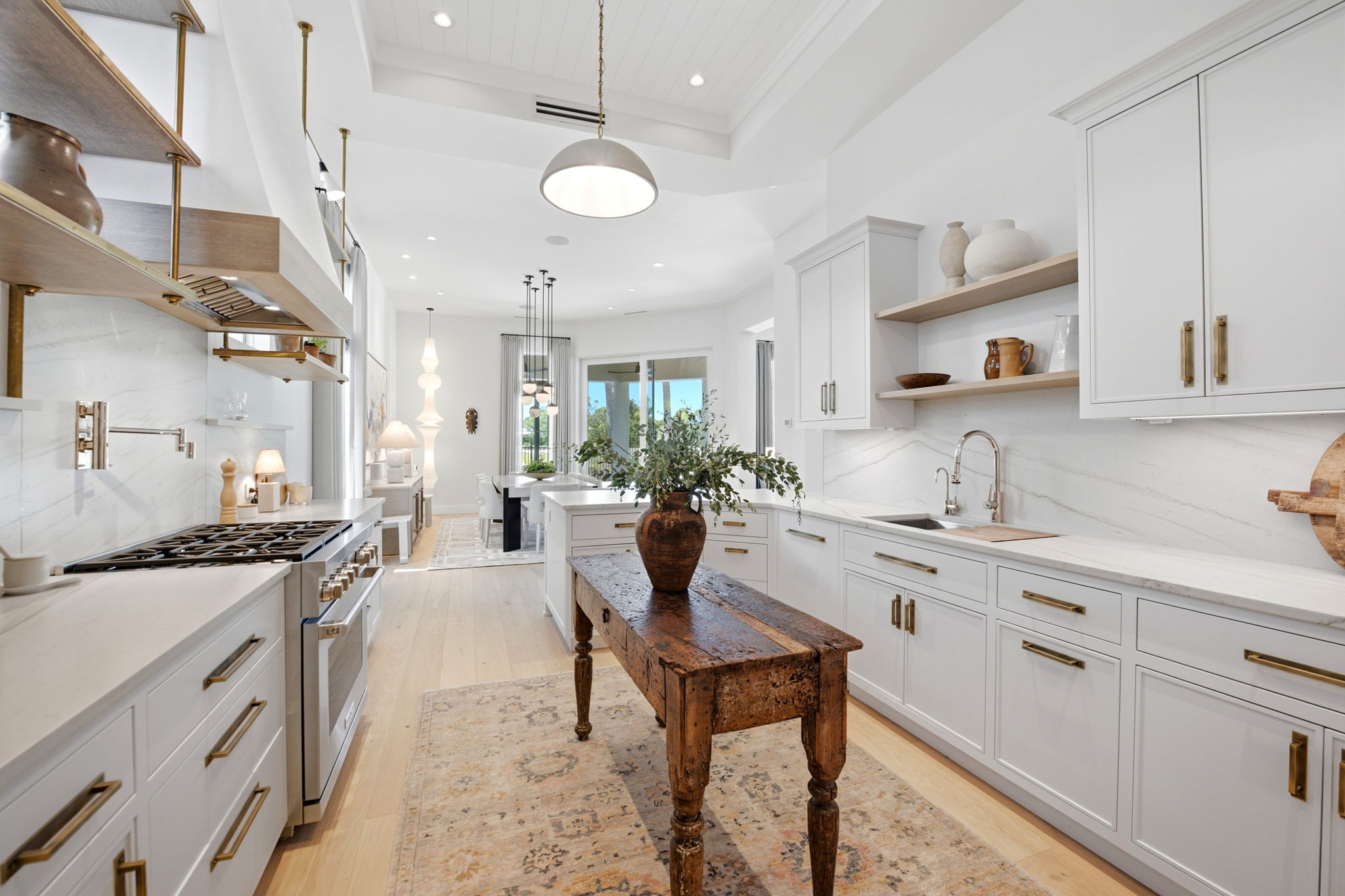 Bright modern kitchen with white cabinetry and brass handles, a rustic wooden table with a large vase of greenery in the center, light wood floors, marble backsplash, and a dining area with pendant lights visible in the background.