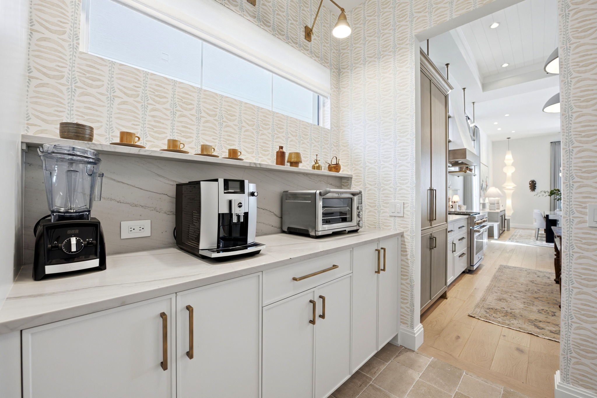 Bright kitchen area with white cabinetry and brass handles, featuring a black blender, silver coffee machine, and stainless steel toaster oven on a marble countertop. Beige patterned wallpaper and a high horizontal window with a white shade provide light. The kitchen opens to a dining area with wood floors, a large rug, tall wooden cabinets, and a modern light fixture in the background.