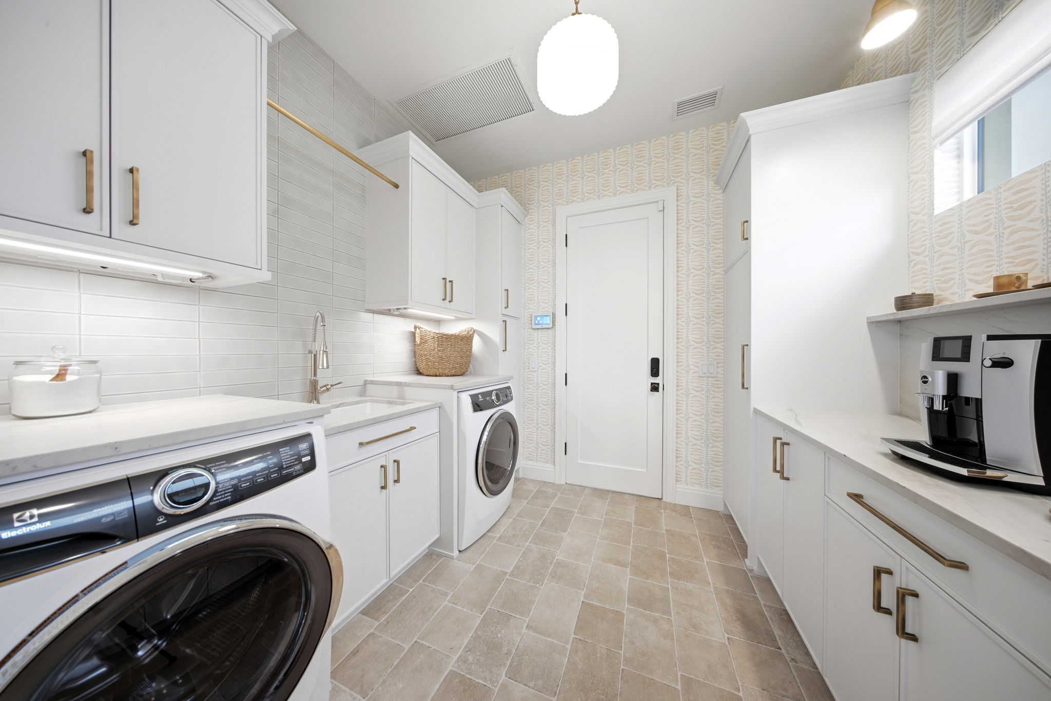 Bright laundry room with beige tiled floor, white cabinetry with brass handles, a front-loading washer and dryer, a white sink with a stainless steel faucet, a wicker basket on the counter, and a coffee machine on the right countertop under a window.