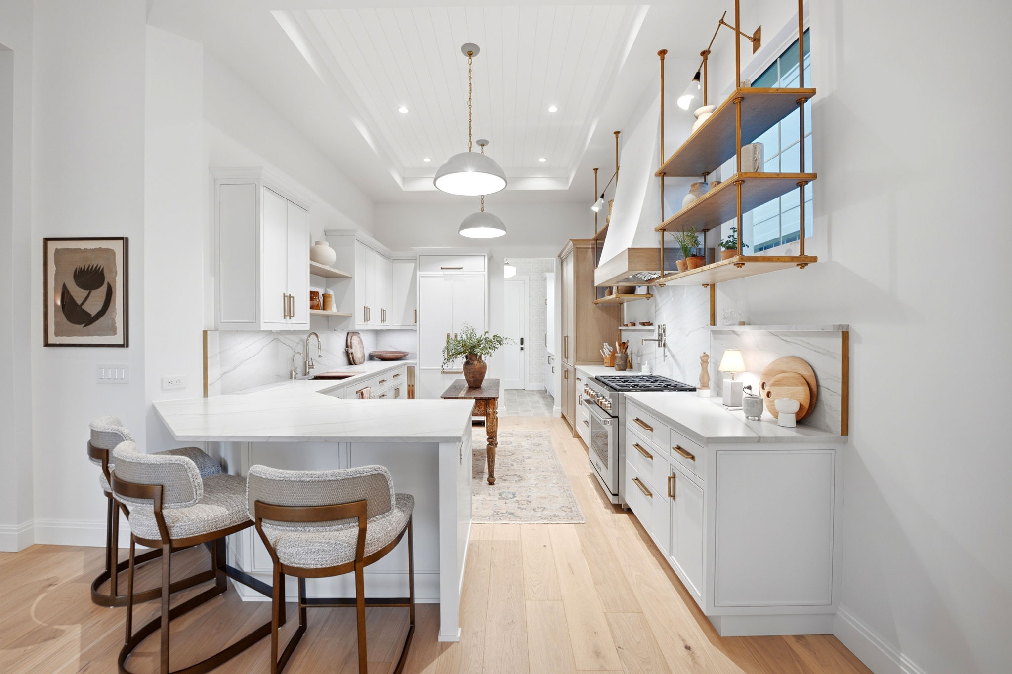 Modern white kitchen with light wooden floor, large island with marble countertop and three cushioned stools, open wooden shelves with plants and kitchenware, stainless steel stove, and a rustic wooden table with a vase of greenery in the center.