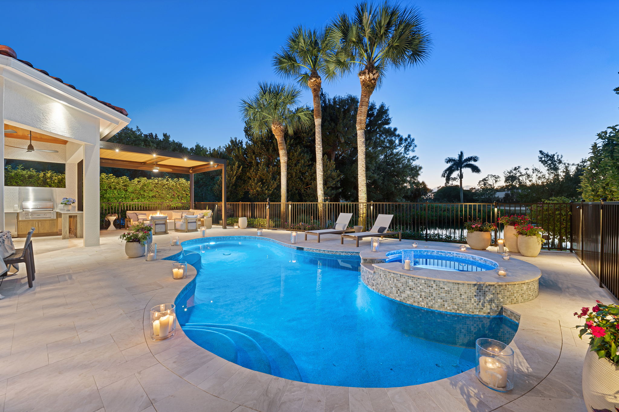 Luxury backyard featuring a lit swimming pool with an attached hot tub surrounded by beige tiled patio, candles in glass holders, potted plants, two lounge chairs, and a covered outdoor seating area with a grill, set against palm trees and a dusk sky.