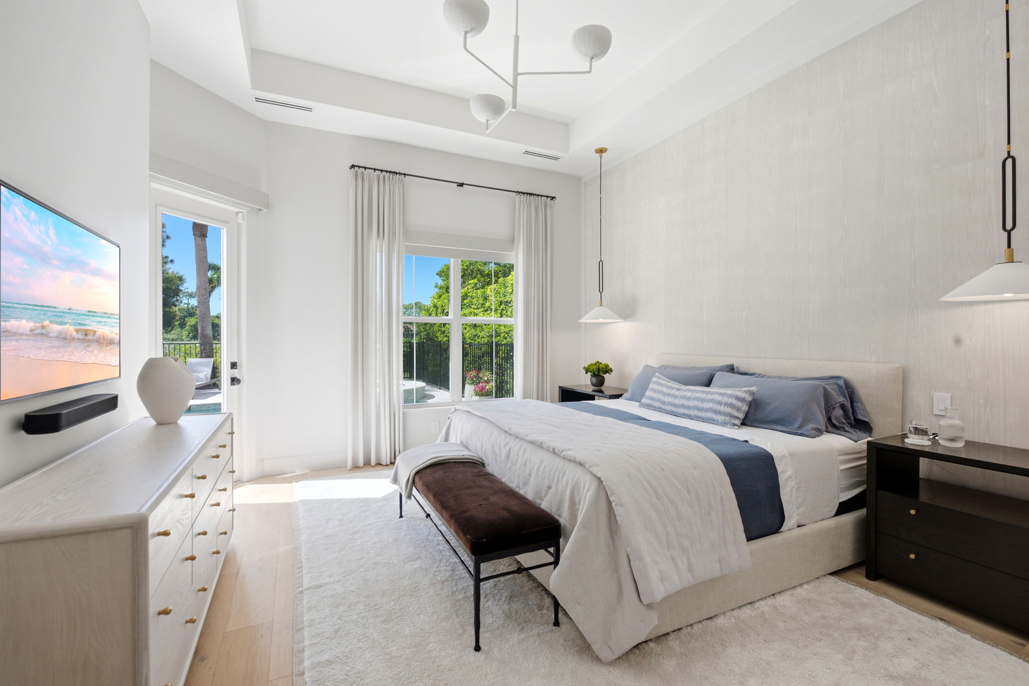 Bright bedroom with a large bed dressed in white and blue bedding, flanked by dark wooden nightstands with hanging pendant lights, a brown bench at the foot of the bed, white dresser with brass knobs, large window, glass door leading outside, and light wood floors with a cream rug.