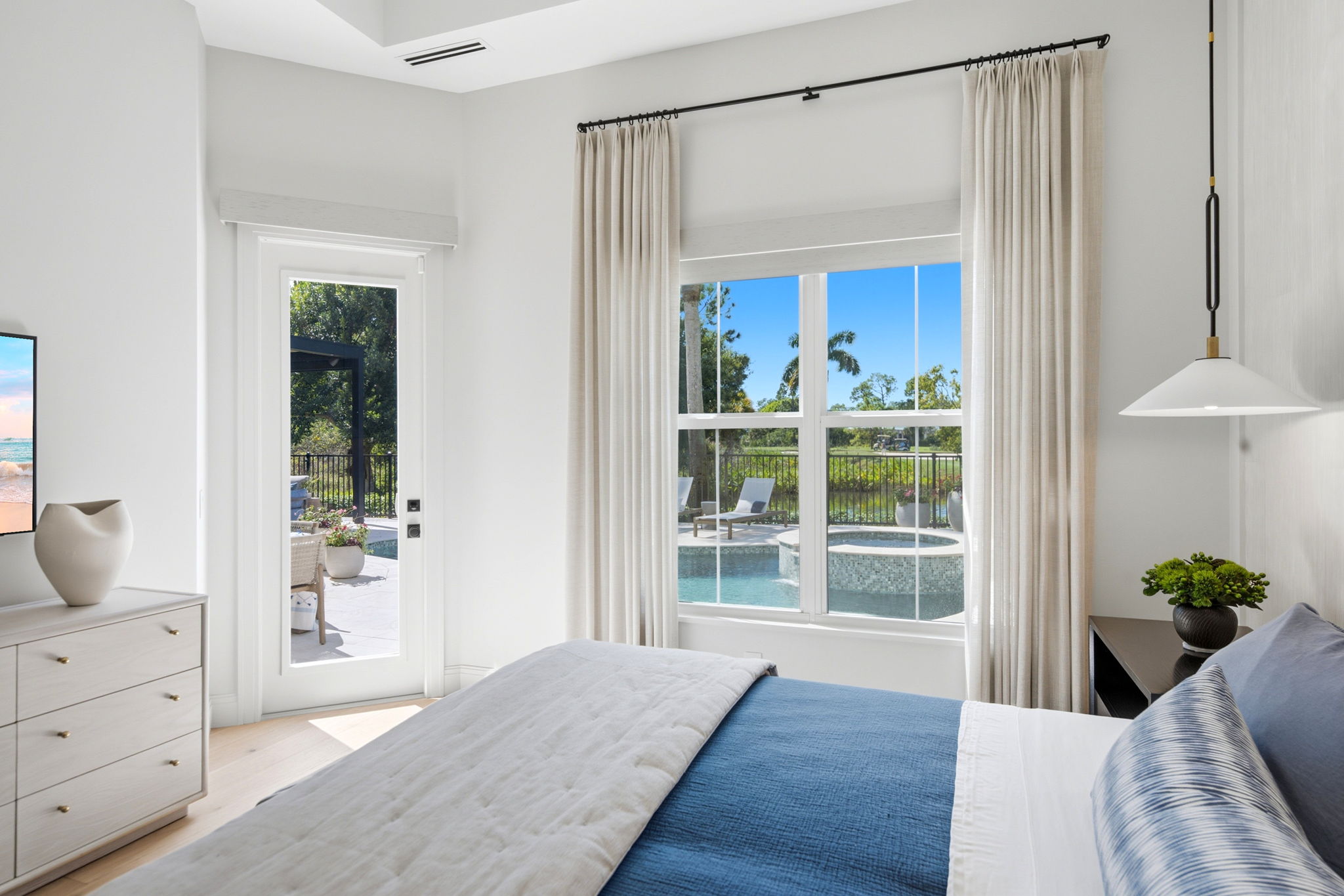 Bright bedroom with white walls, a large window showing a pool and palm trees outside, a door leading to the patio, a bed dressed in blue and white bedding, a dresser with a white vase, and a nightstand with a green plant and modern pendant light.