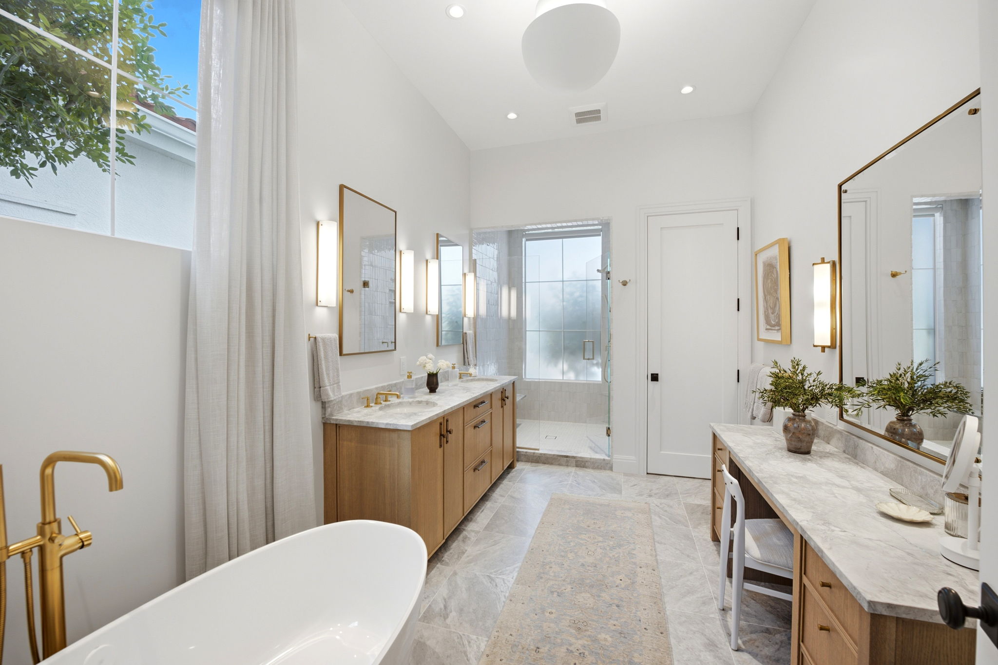 Bright modern bathroom with a freestanding white tub and gold faucet near a large window with sheer curtains, a double vanity with marble countertop, gold hardware, two mirrors with vertical lights, a walk-in shower with frosted glass window, and a large vanity desk with a matching marble top, chair, and vase with greenery.