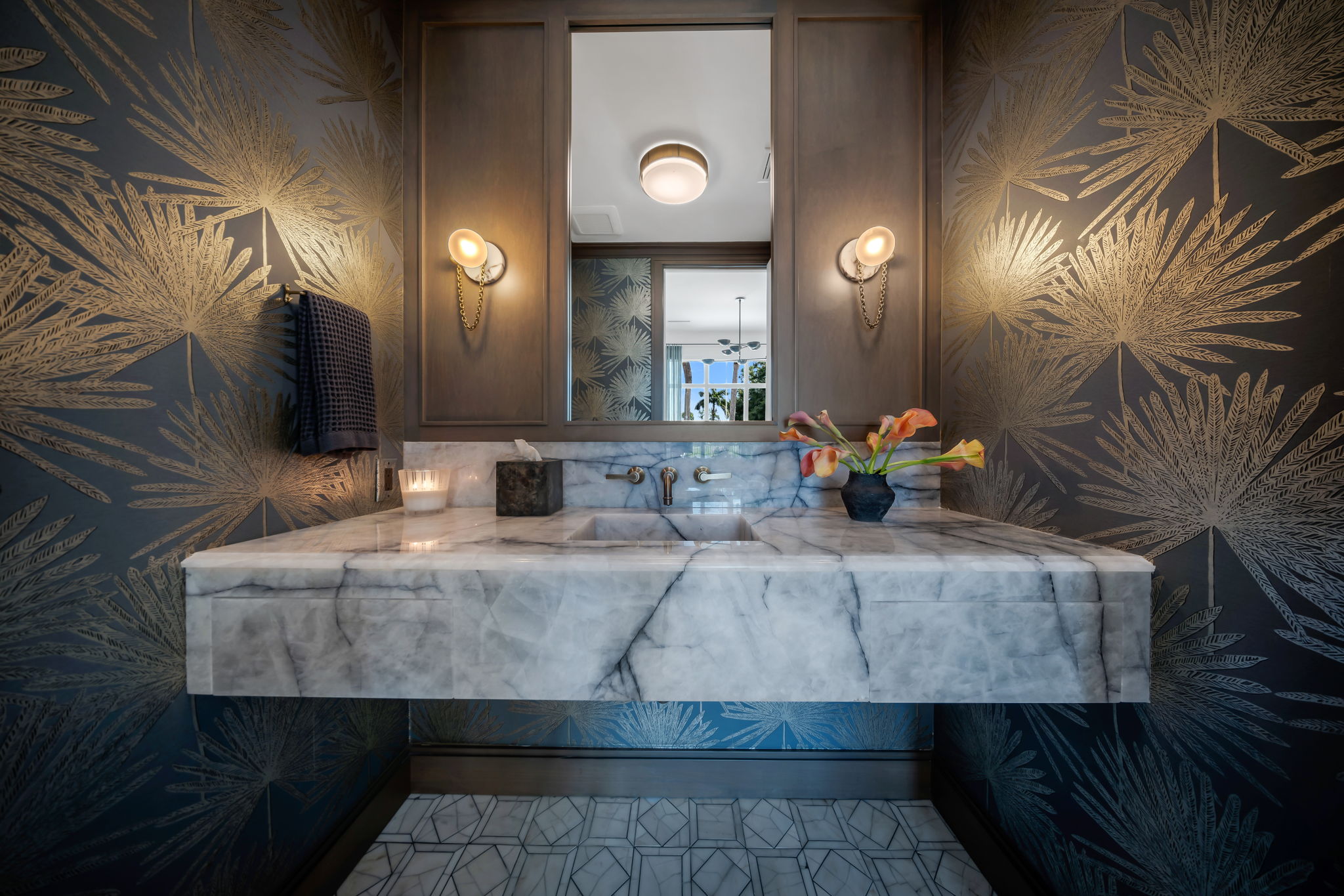 Modern bathroom sink area with floating white marble countertop and integrated rectangular basin, flanked by two gold wall sconces with chain details, set against wallpaper with large gold leaf patterns on dark background, a dark towel hanging on the left, a lit candle and tissue box on the counter, and a small vase holding orange calla lilies on the right, with a large mirror reflecting a window and ceiling light.