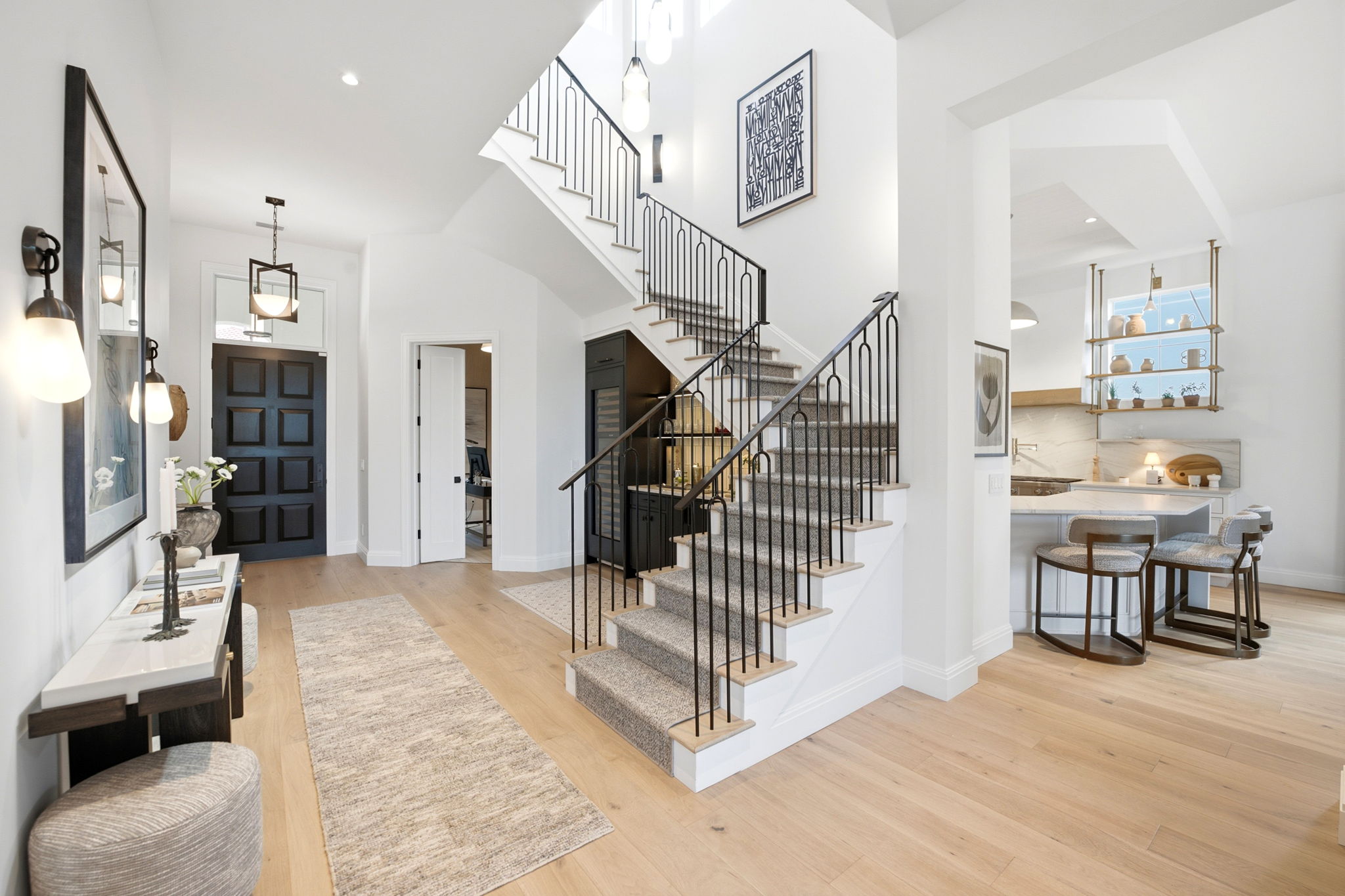 Modern home's bright entryway with light wood flooring, black front door, staircase with metal railing and carpet runner, console table with decor, and open view into kitchen with bar stools and shelves.