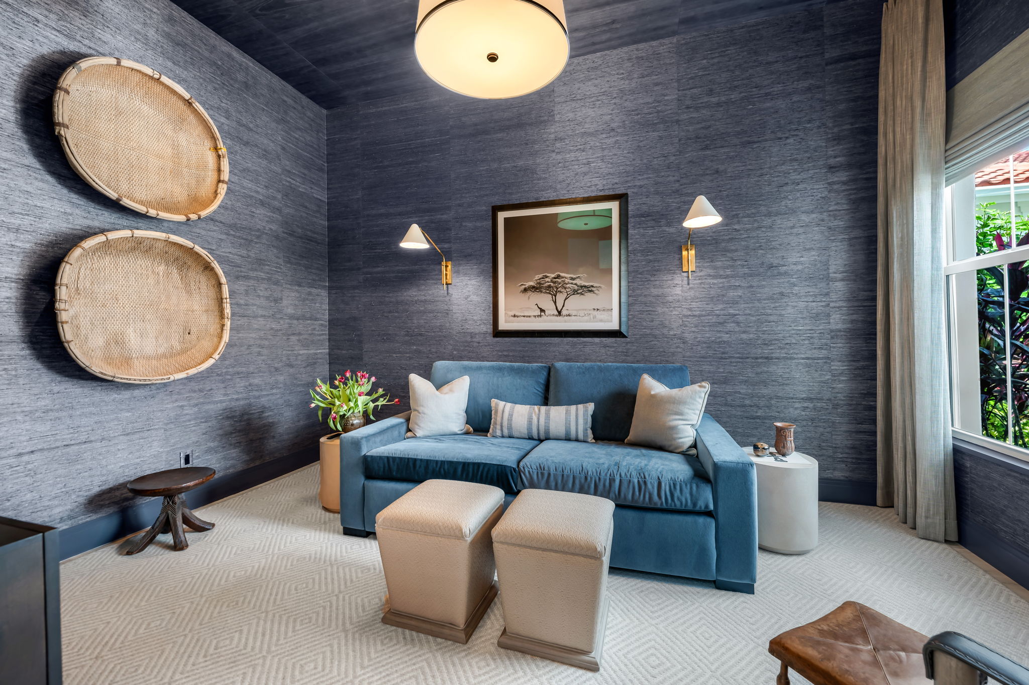 Cozy living room with a blue sofa adorned with three cushions, two small beige ottomans, a wooden stool, and a light beige patterned carpet. Two woven baskets hang on the dark textured walls. Above the sofa is a framed black-and-white picture of a tree and giraffes. Two wall lamps flank the picture. A side table with a vase of pink tulips is next to the sofa, and another small table with decorative items sits by a window with light curtains, showing greenery outside.