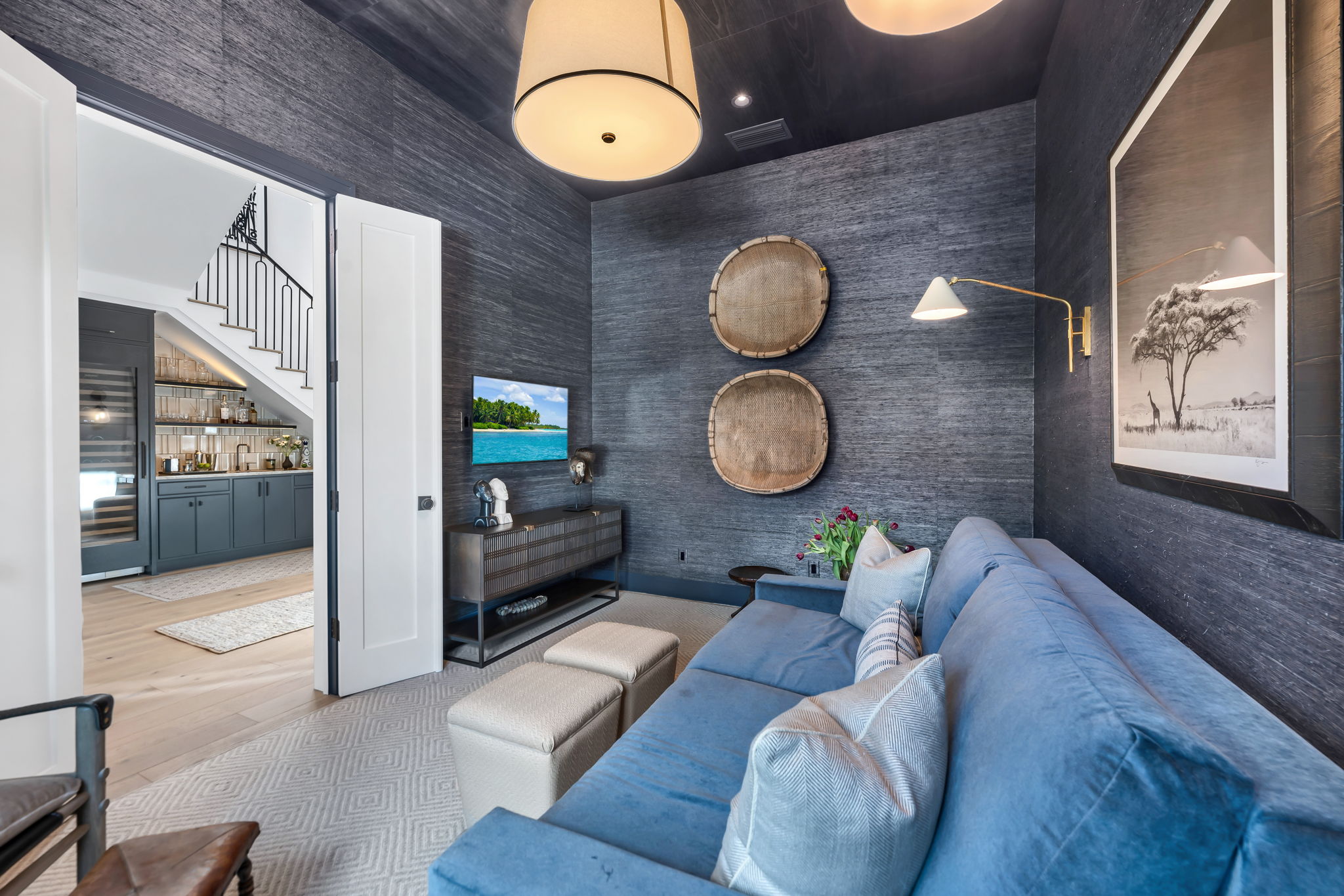 Cozy living room with dark textured walls, a large blue sofa with white pillows, two beige ottomans, and a wall-mounted TV showing a tropical island scene; decorative baskets hang on the wall above a modern console table with sculptures; an open door reveals a kitchen area with dark cabinetry, shelves, and a staircase.