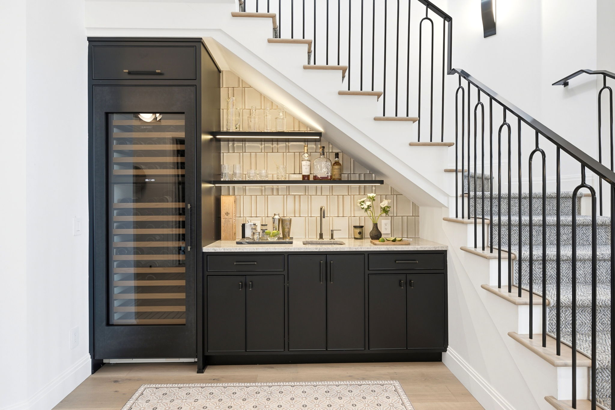 Modern under-stair bar area with black cabinets, a wine fridge on the left, a small sink, and two illuminated shelves holding glassware and bottles, set against a beige tiled backsplash.
