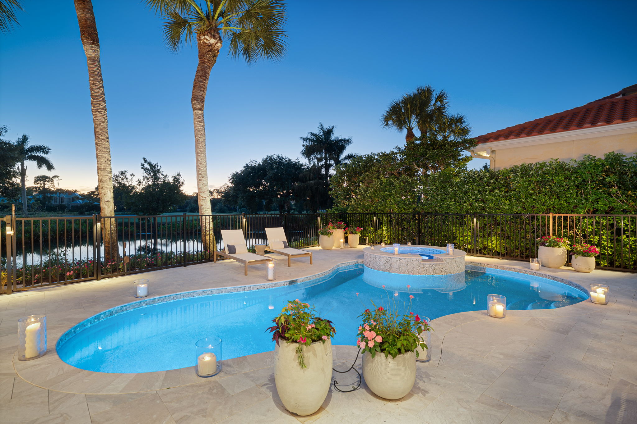 Outdoor swimming pool at dusk with built-in hot tub, surrounded by tiled deck, potted plants, lit candles, two lounge chairs, palm trees, and a black metal fence overlooking a body of water and greenery.