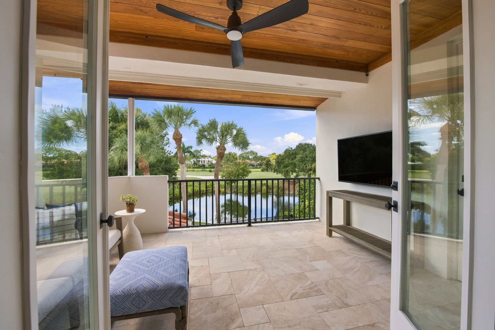 Covered balcony with beige tiled floor, wooden ceiling, and black ceiling fan, furnished with a blue patterned ottoman, small white side table with a plant, and a wall-mounted TV, overlooking palm trees, pond, and landscaped golf course under a clear blue sky.