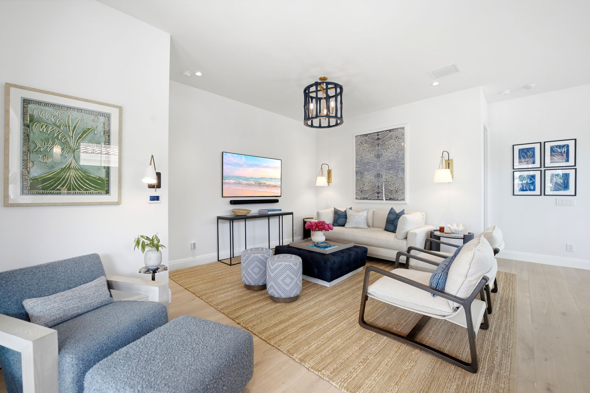 Bright modern living room with white walls, a beige sofa with blue pillows, two beige armchairs with dark wooden frames, a blue armchair with matching ottoman, a navy tufted ottoman coffee table with a vase of pink flowers, two patterned round ottomans, a wall-mounted TV above a narrow black console table, and framed abstract and botanical artwork on the walls.
