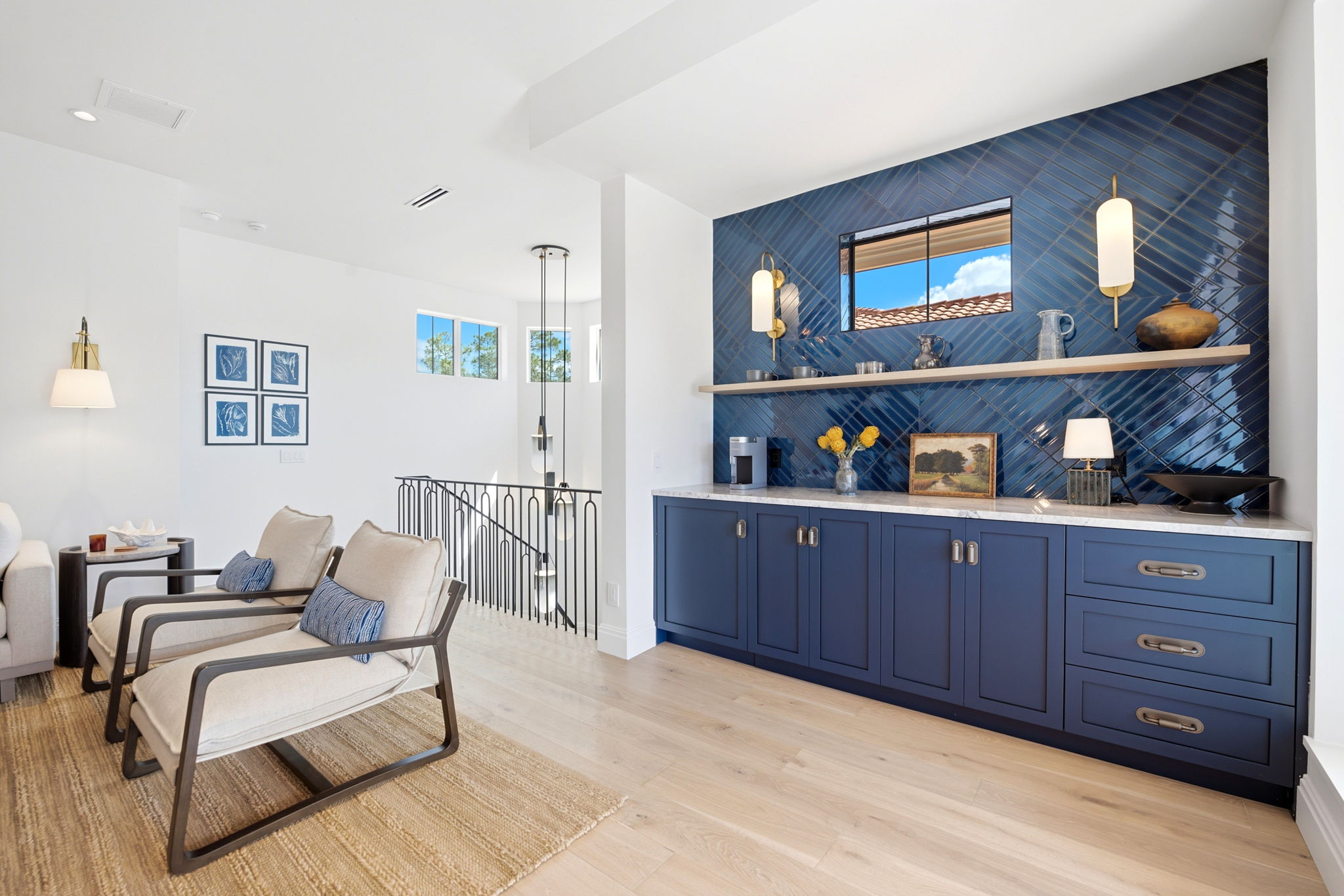 Modern living space with two beige cushioned armchairs with dark frames on a woven rug, next to a blue cabinet with a white countertop and blue tile backsplash; decorations include a coffee maker, vases, a framed landscape painting, and brass and white wall sconces, with a window reflecting a blue sky and roof tiles.