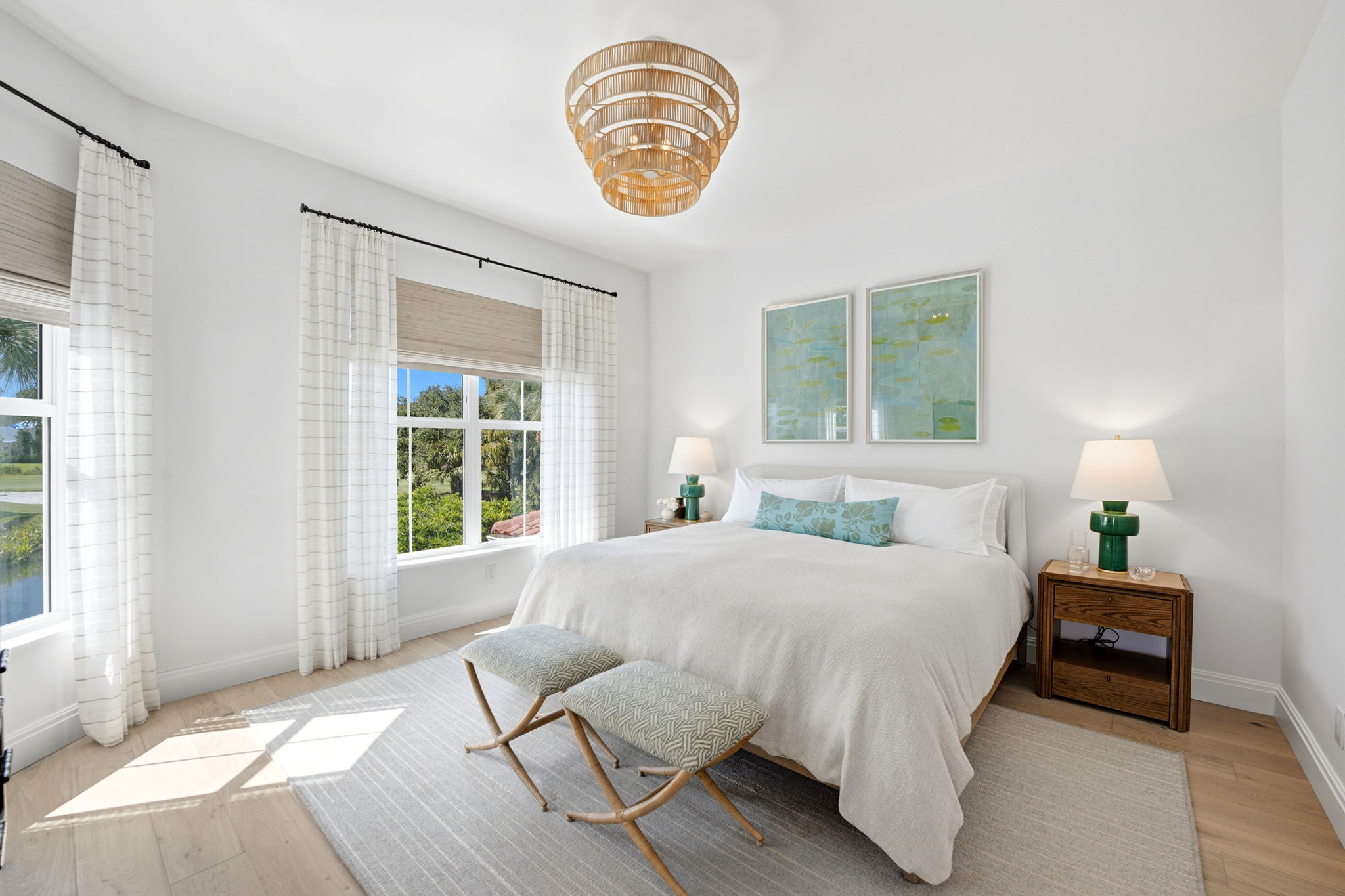 Bright bedroom with white walls, large bed with white bedding and blue accent pillow, two light blue patterned stools at the foot of the bed, wooden nightstands with green lamps, two framed botanical prints above the bed, large windows with white curtains letting in natural light, and light wood flooring.
