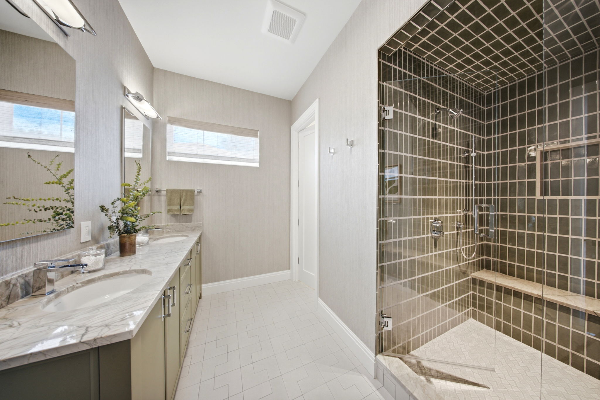 Bright modern bathroom with double sinks in marble countertop, large mirror, small window above, and a glass-enclosed tiled shower with bench seat and chrome fixtures.