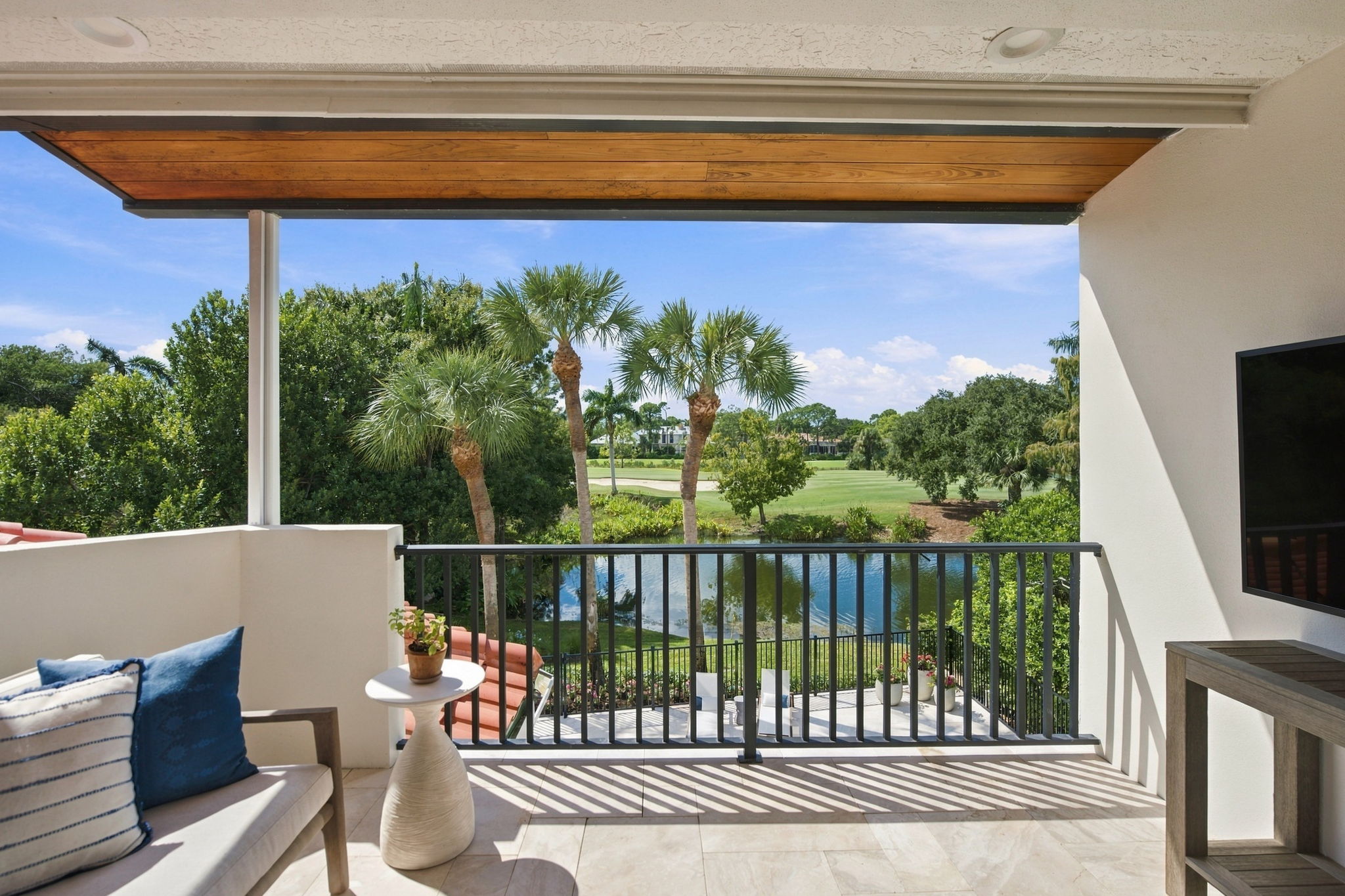 Covered balcony with gray metal railing overlooking a pond, palm trees, and a golf course under a partly cloudy sky, featuring a cushioned bench with blue and white pillows, a small side table with a potted plant, and a wall-mounted TV.