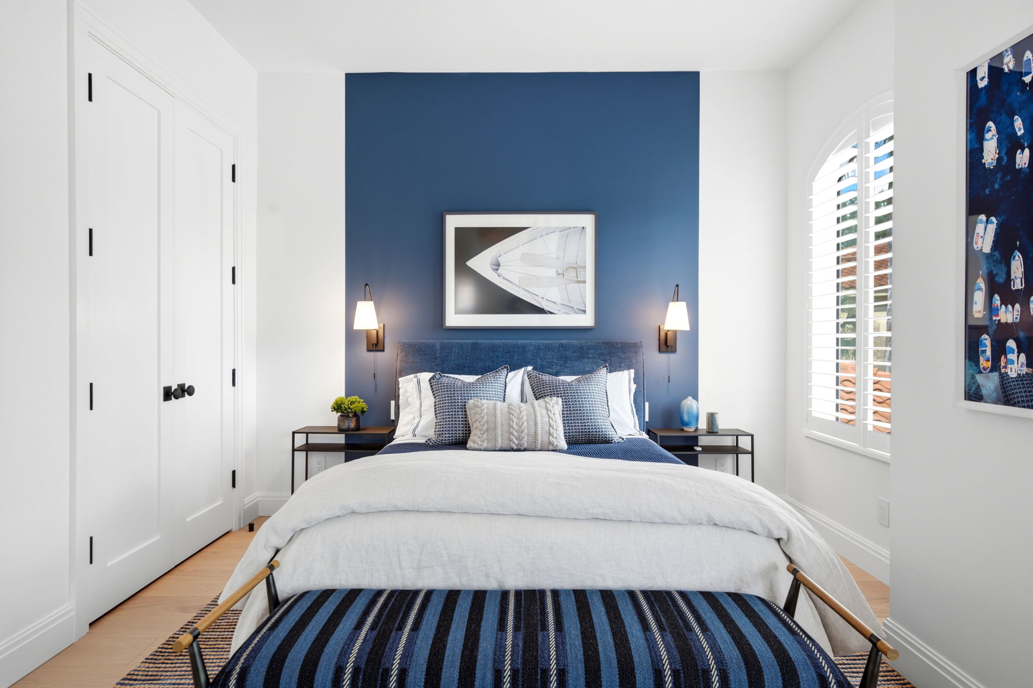 Contemporary bedroom with a blue accent wall behind a bed with a blue upholstered headboard, blue and white pillows, white bedding, and a striped blue bench at the foot, flanked by two black nightstands with lamps and decor, a large window with shutters on the right, and a white closet door on the left.