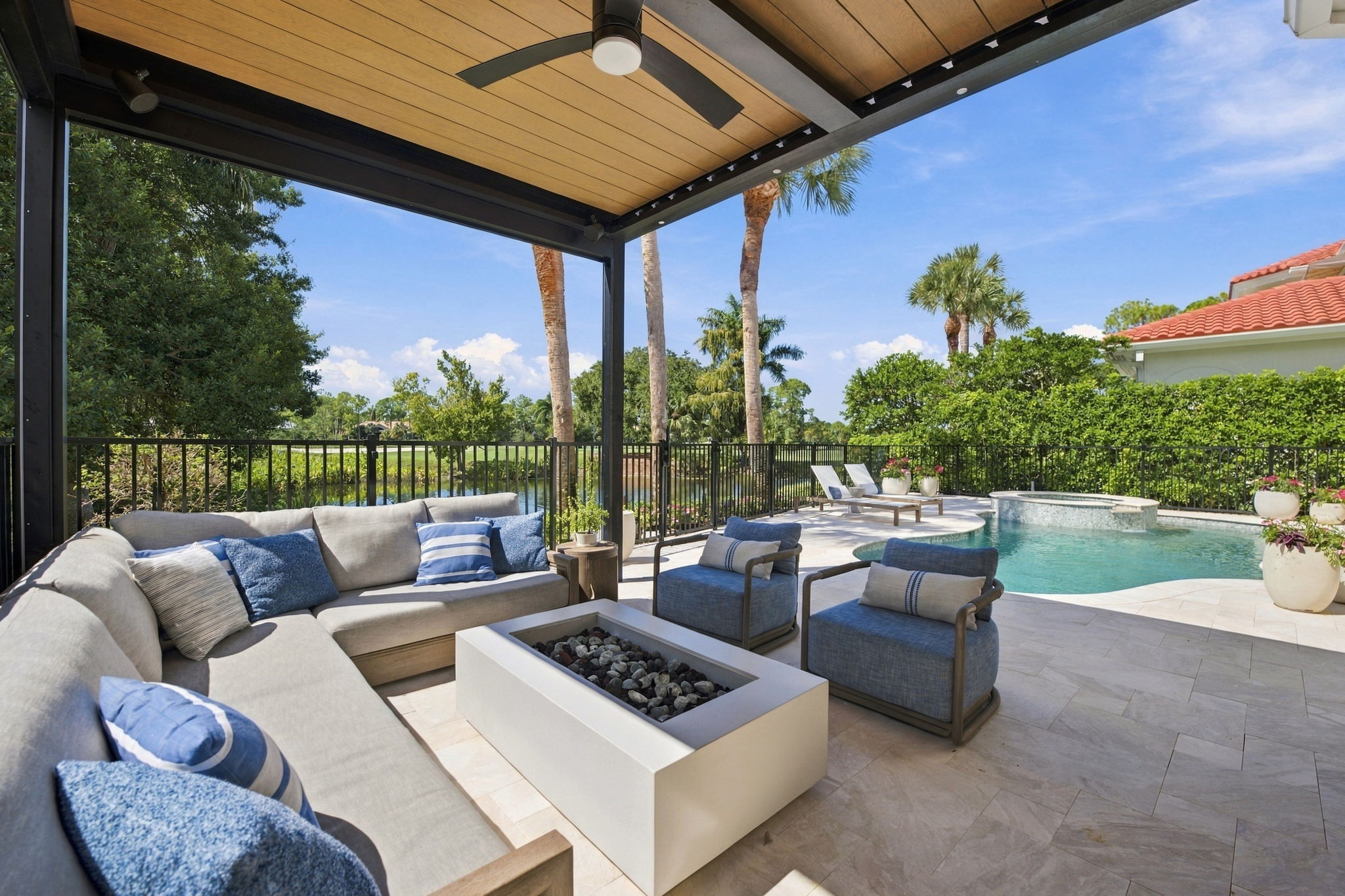 Outdoor patio with a covered seating area featuring a gray sectional sofa with blue and striped cushions, two blue armchairs with matching pillows, and a rectangular fire pit table, overlooking a fenced swimming pool and lush green landscaping with palm trees under a clear blue sky.