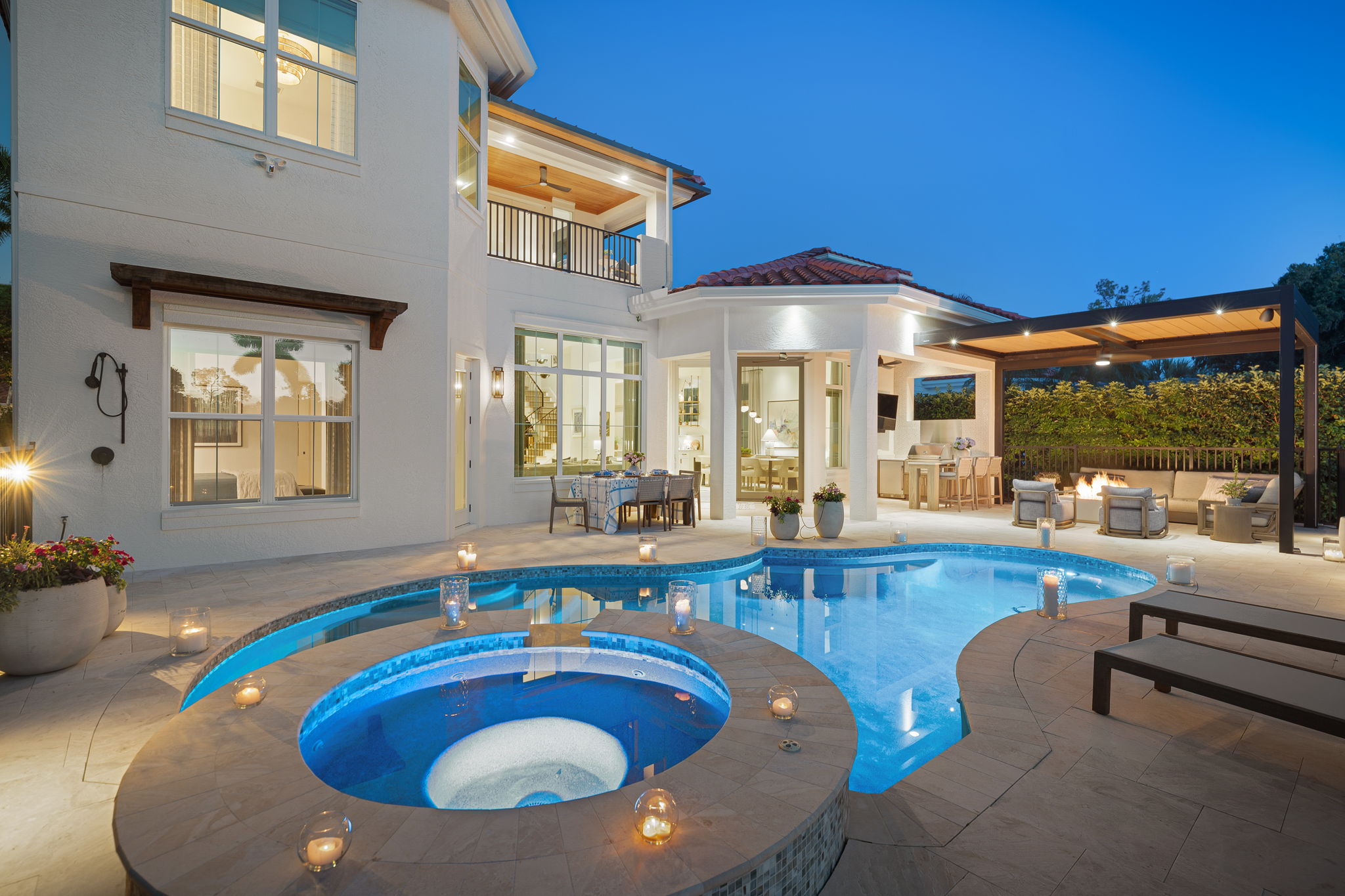Luxury backyard at dusk featuring a lit swimming pool with an attached circular hot tub surrounded by small candles, outdoor dining table, covered seating area with firepit, and a modern two-story white house with large windows and balcony.