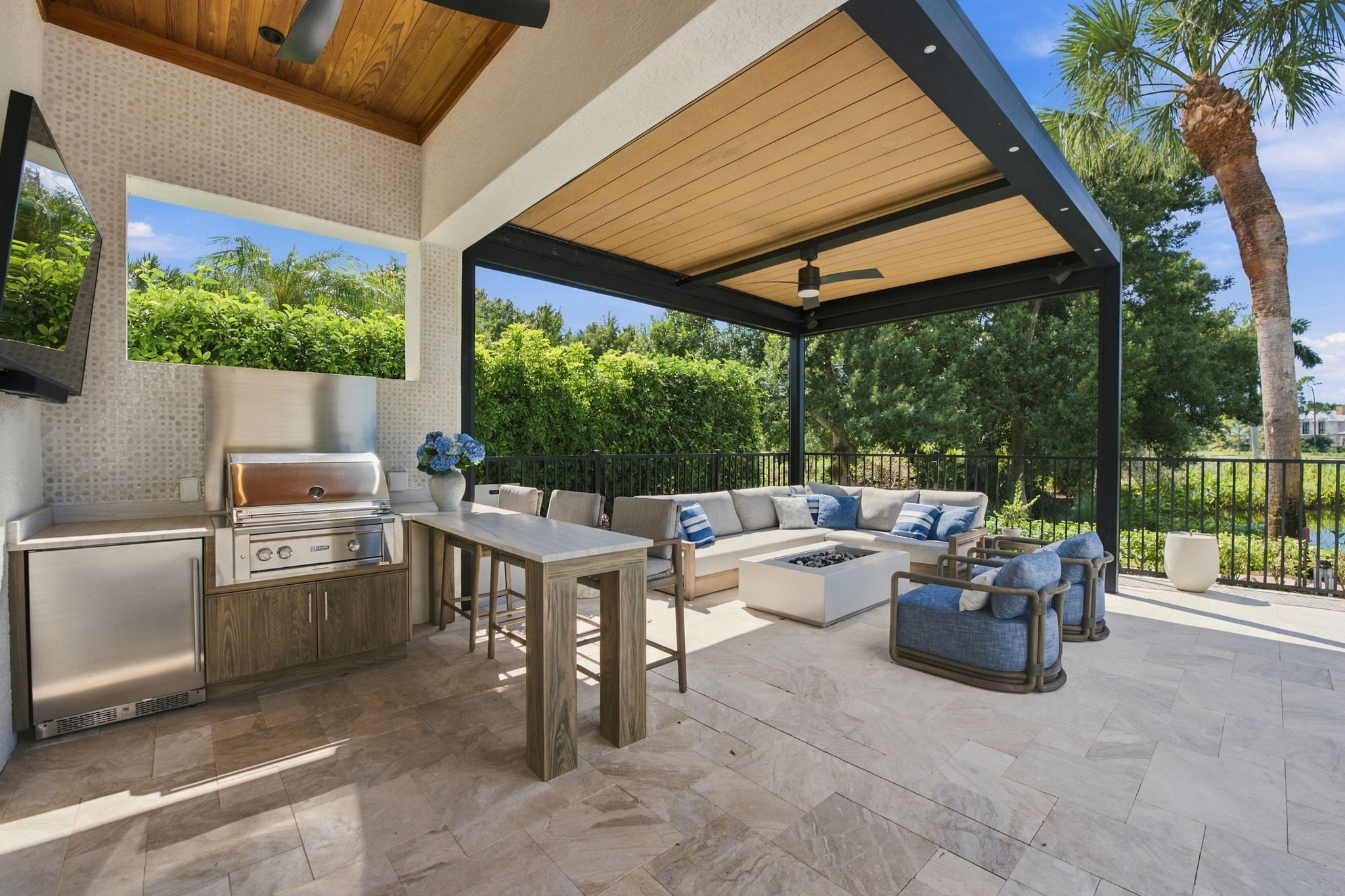 Outdoor covered patio featuring a stainless steel built-in grill with counter space and a small fridge, a tall wooden table with two chairs, and a seating area with a large L-shaped sofa, two blue cushioned armchairs, a square fire pit, and ceiling fan under a wooden panel roof, surrounded by lush greenery and palm trees.