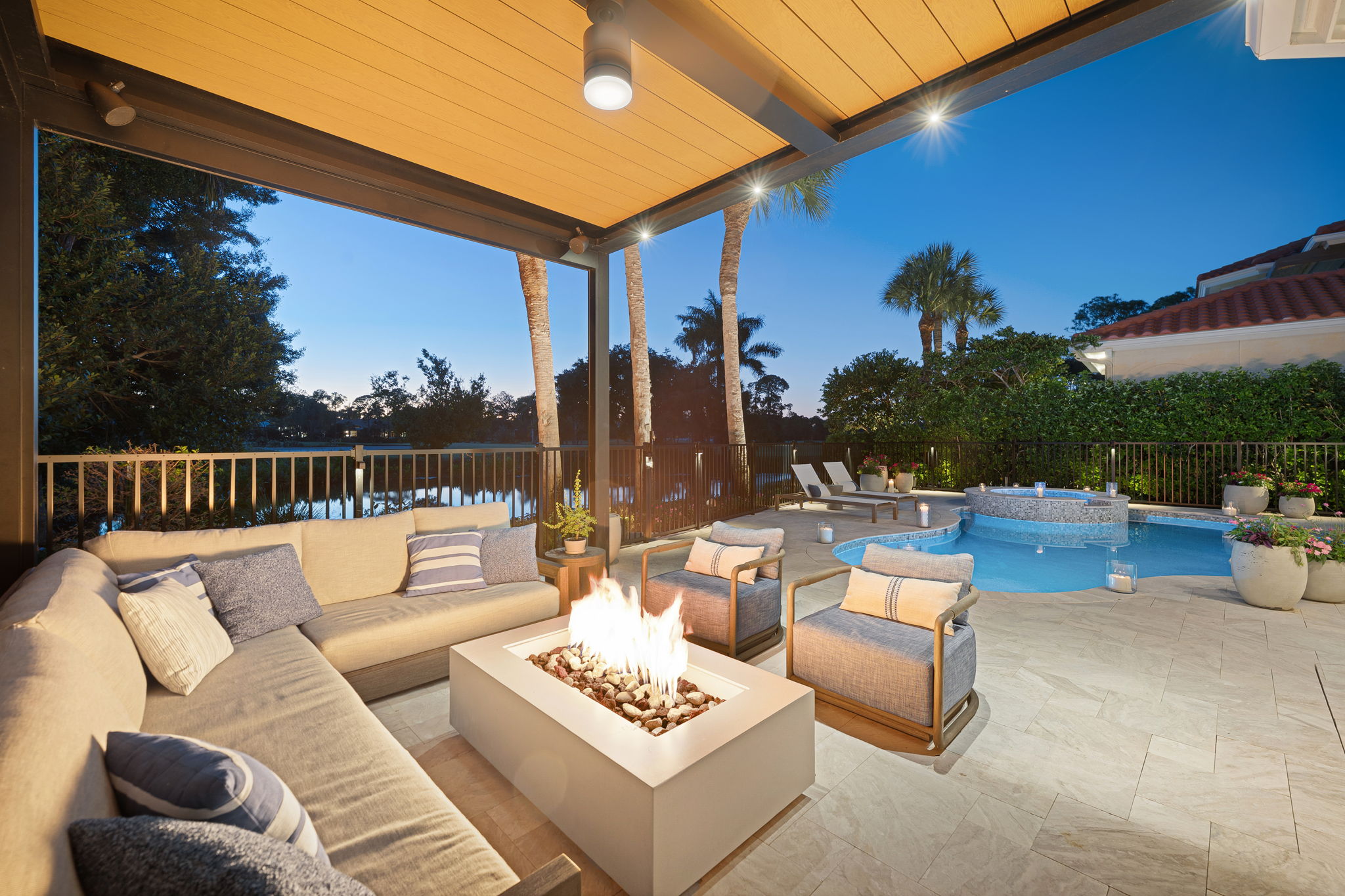 Outdoor patio at dusk with a beige sectional sofa and two cushioned chairs around a rectangular fire pit, under a wooden ceiling, overlooking a pool with a hot tub, surrounded by lounge chairs, potted plants, and palm trees.