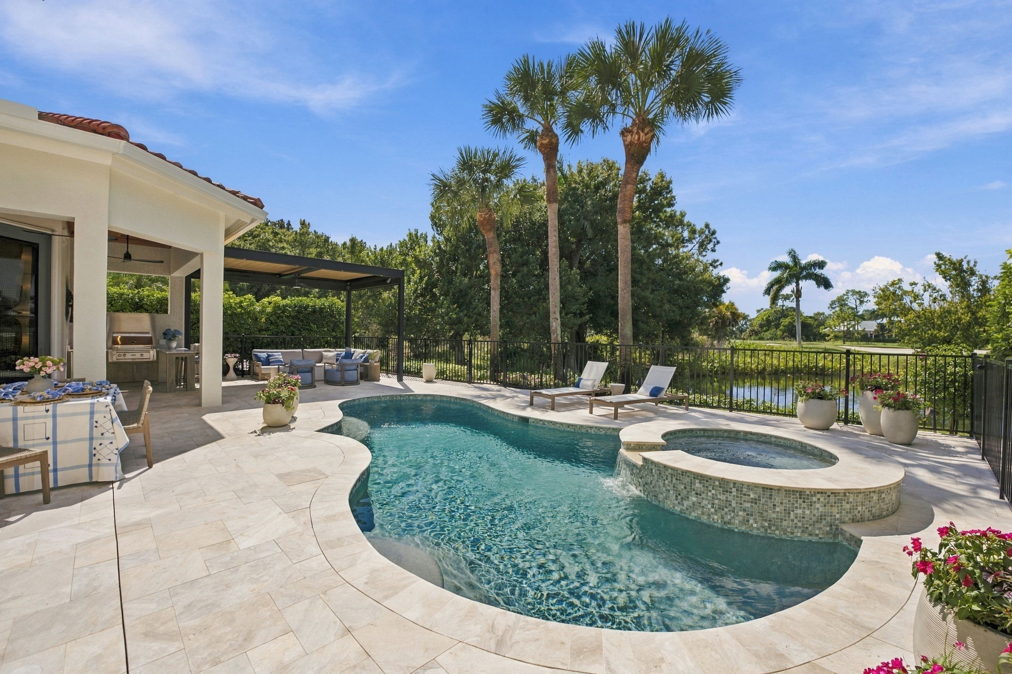 Backyard with a curved swimming pool connected to a round hot tub, surrounded by beige tile decking, three palm trees, potted plants, two lounge chairs, an outdoor seating area with a pergola, and a dining table with a blue and white tablecloth under clear blue sky.