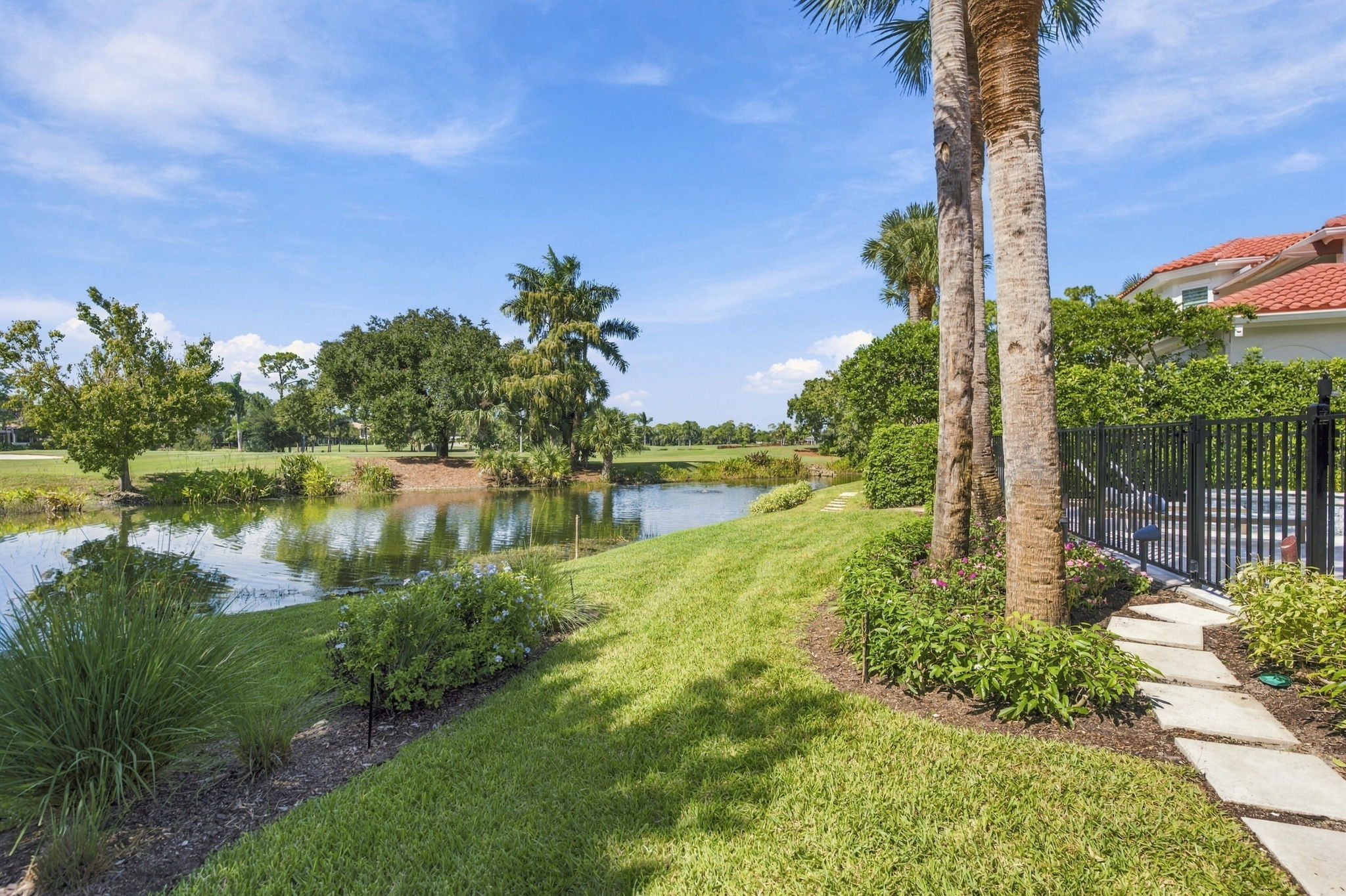 A lush green lawn beside a calm pond with reflections of trees under a blue sky, a path with stepping stones leads to a black metal fence near palm trees and a house with a red-tiled roof.