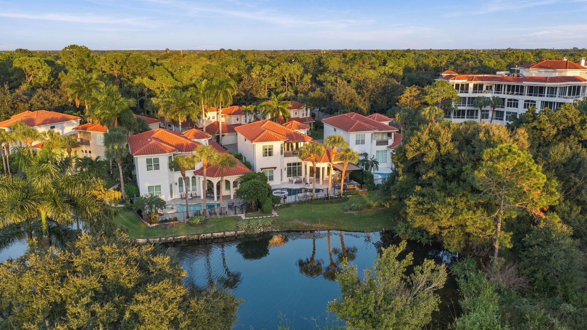 Aerial view of several upscale houses with red-tiled roofs and palm trees surrounding a small pond, set against a backdrop of a dense green forest under a partly cloudy sky.