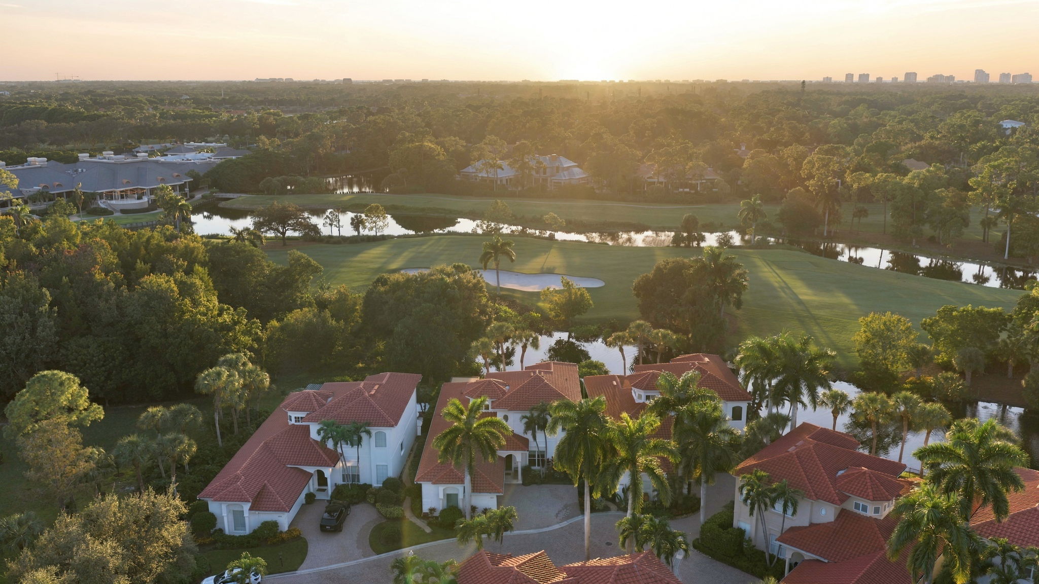 Aerial view at sunset of a residential area with large houses featuring red tiled roofs, surrounded by palm trees and greenery, adjacent to a golf course with sand traps and water hazards, under a clear sky.