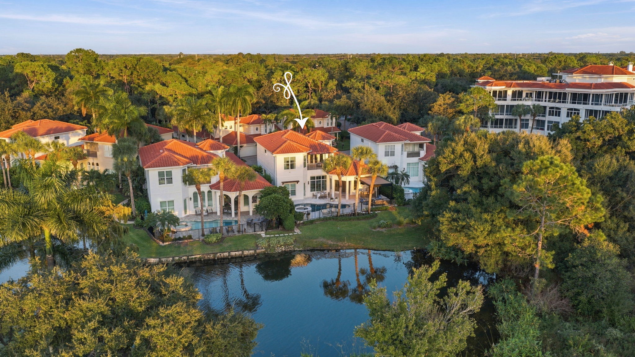 Aerial view of a residential neighborhood with Mediterranean-style houses with red tile roofs surrounded by lush greenery and palm trees, centered on a two-story house marked with a decorative white arrow. In the foreground is a calm water body reflecting trees and homes.