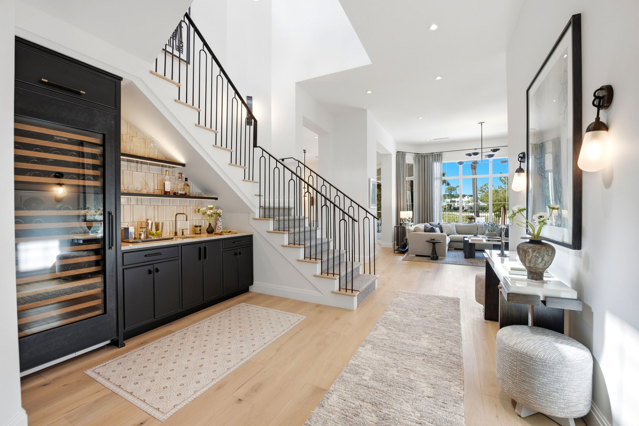 Modern open living space with light wood flooring, stairway with black metal railing and carpet runner, built-in dark cabinetry with wine fridge under stairs, and a bright living room featuring a large window with garden view, neutral sofas, and sheer curtains.