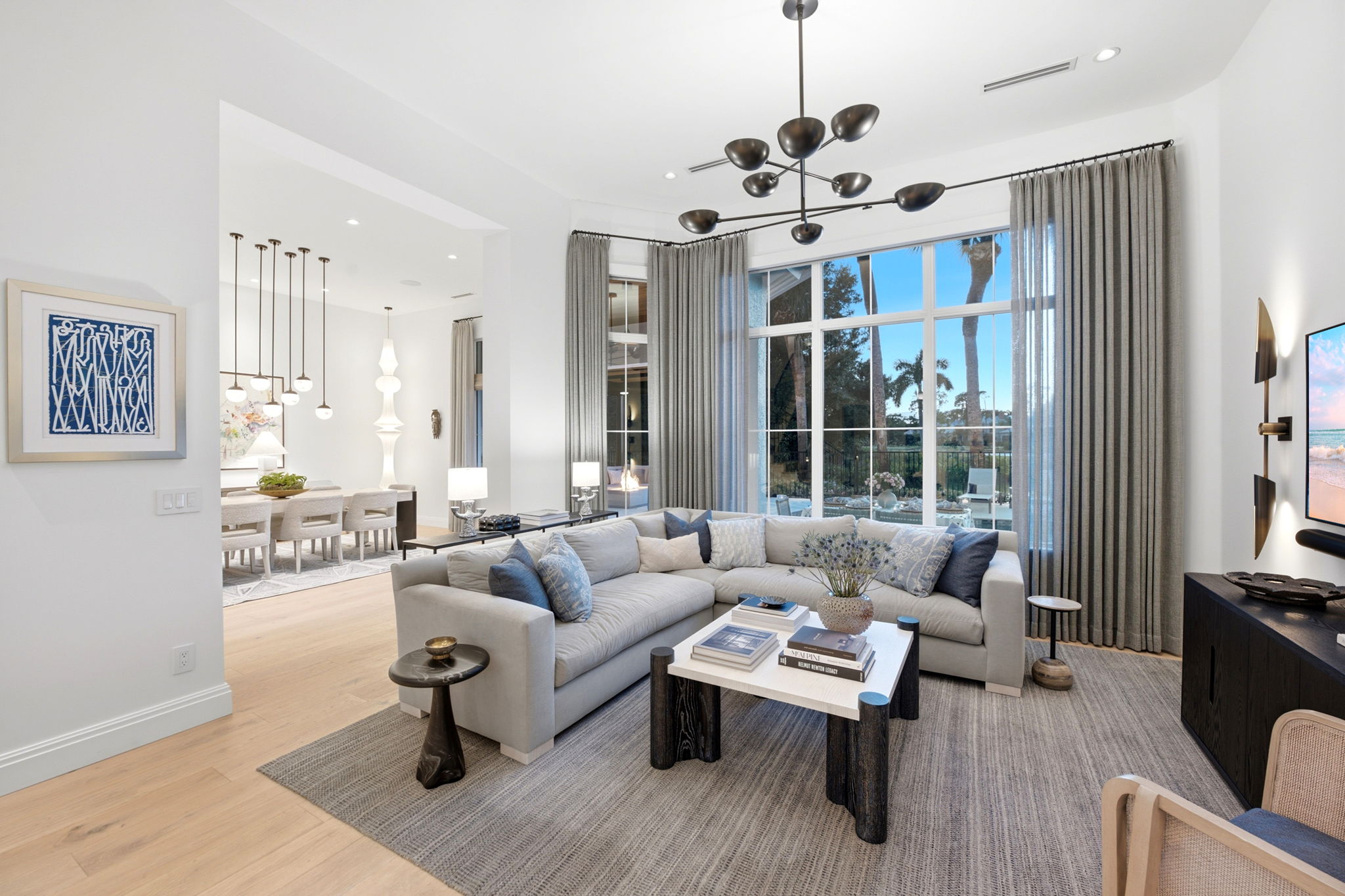 Bright modern living room with a large light gray sectional sofa adorned with blue and gray pillows, a white-top coffee table with black legs holding books and a vase with flowers, a small black side table, and a gray area rug. Floor-to-ceiling windows with gray curtains show outdoor palm trees. A wall-mounted TV is on the right, and a dining area with a white table, chairs, and pendant lighting is visible through an open doorway.