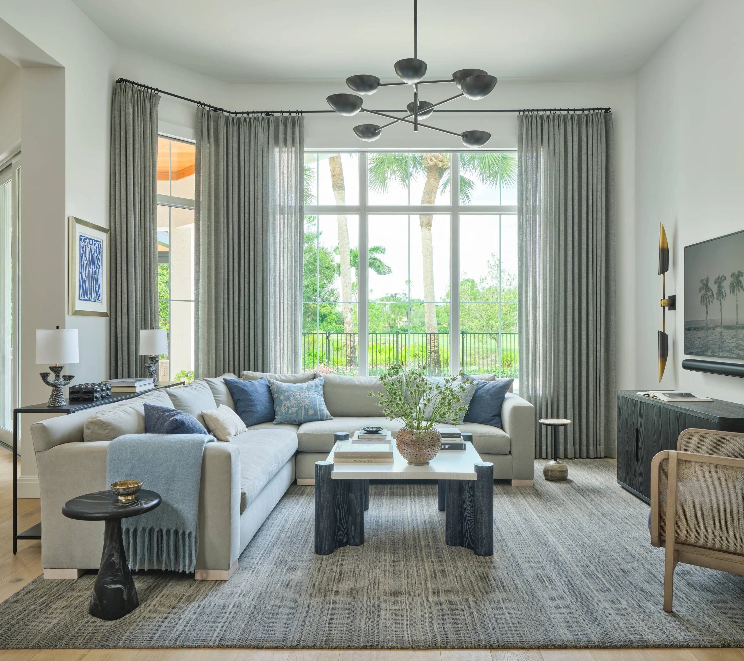Bright living room with a large beige sectional sofa adorned with blue and beige throw pillows, a blue throw blanket, a white and dark wood coffee table with books and a vase with flowers, and a modern black side table with a gold bowl. Floor-to-ceiling windows with sheer gray curtains reveal green trees and palm trees outside. The room also features light wooden floors paired with a large textured gray area rug, a flat-screen TV on a dark wooden console, two table lamps on a black metal console behind the sofa, and a modern black chandelier hanging from the ceiling.