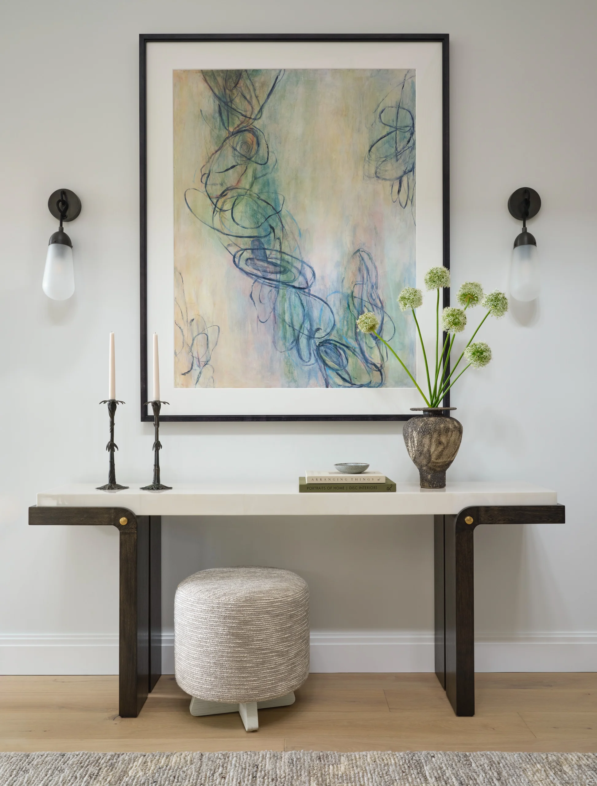 Minimalist entryway with a white and dark wood console table holding two black candle holders with white candles, stacked books, a small dish, and a textured vase with round white flowers; a modern abstract painting in a black frame hangs above, flanked by two black wall sconces with frosted glass shades, a round woven ottoman sits beneath the table on a light wood floor and a beige textured rug.