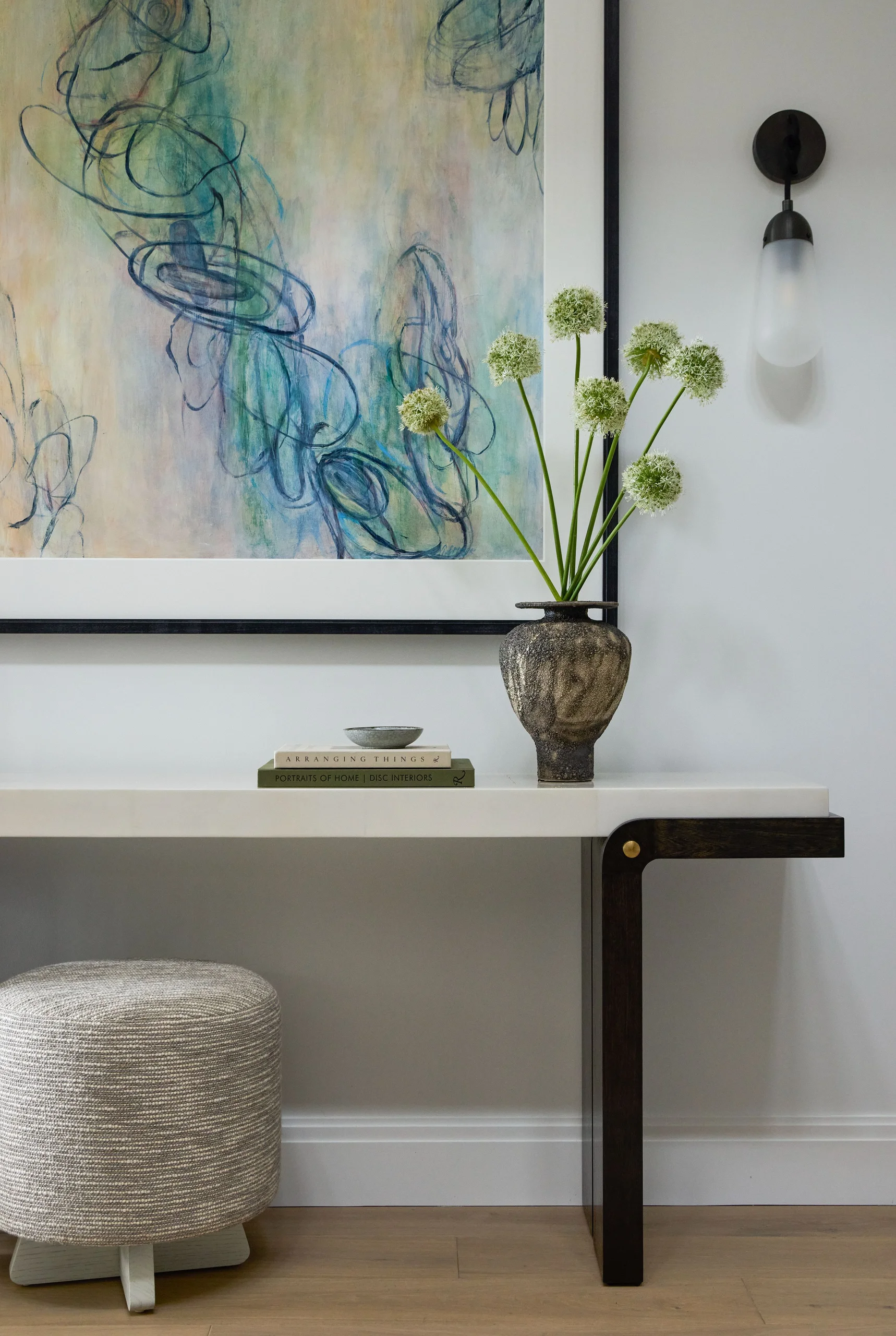 Minimalist interior scene with a marble-top console table featuring dark wood leg accents, topped with a textured ceramic vase holding white spherical flowers, two stacked books, and a small dish. A large abstract painting with blue and beige tones hangs above the table against a white wall, beside a modern wall sconce with a frosted glass shade. A round, woven beige ottoman sits beneath the table on a light wooden floor.