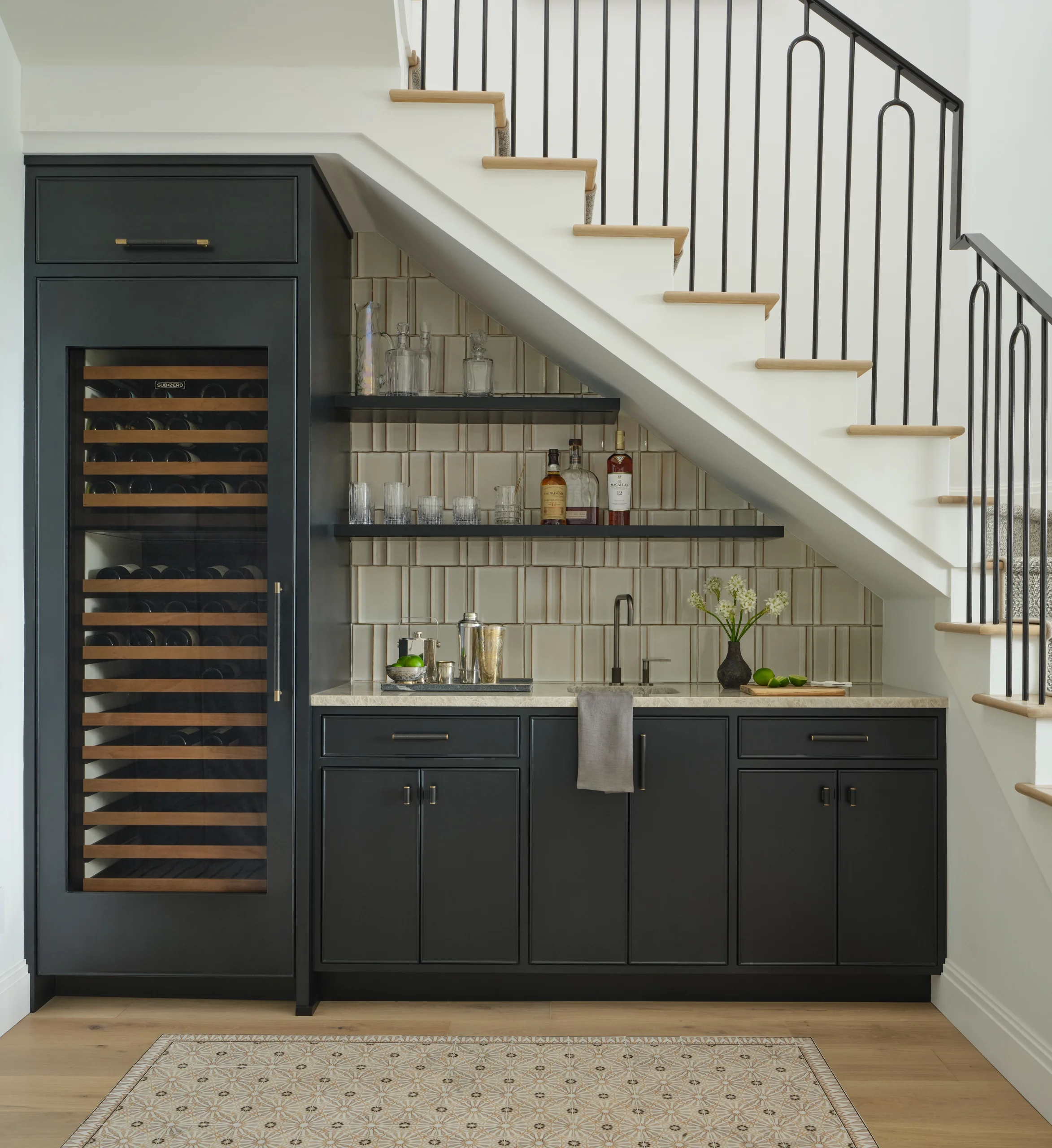 Modern home bar setup under a staircase with black cabinetry, marble countertop, sink with faucet, glass shelves holding bottles and glasses, built-in wine cooler with wooden racks, decorative vase with flowers, and a patterned rug on light wood floor.