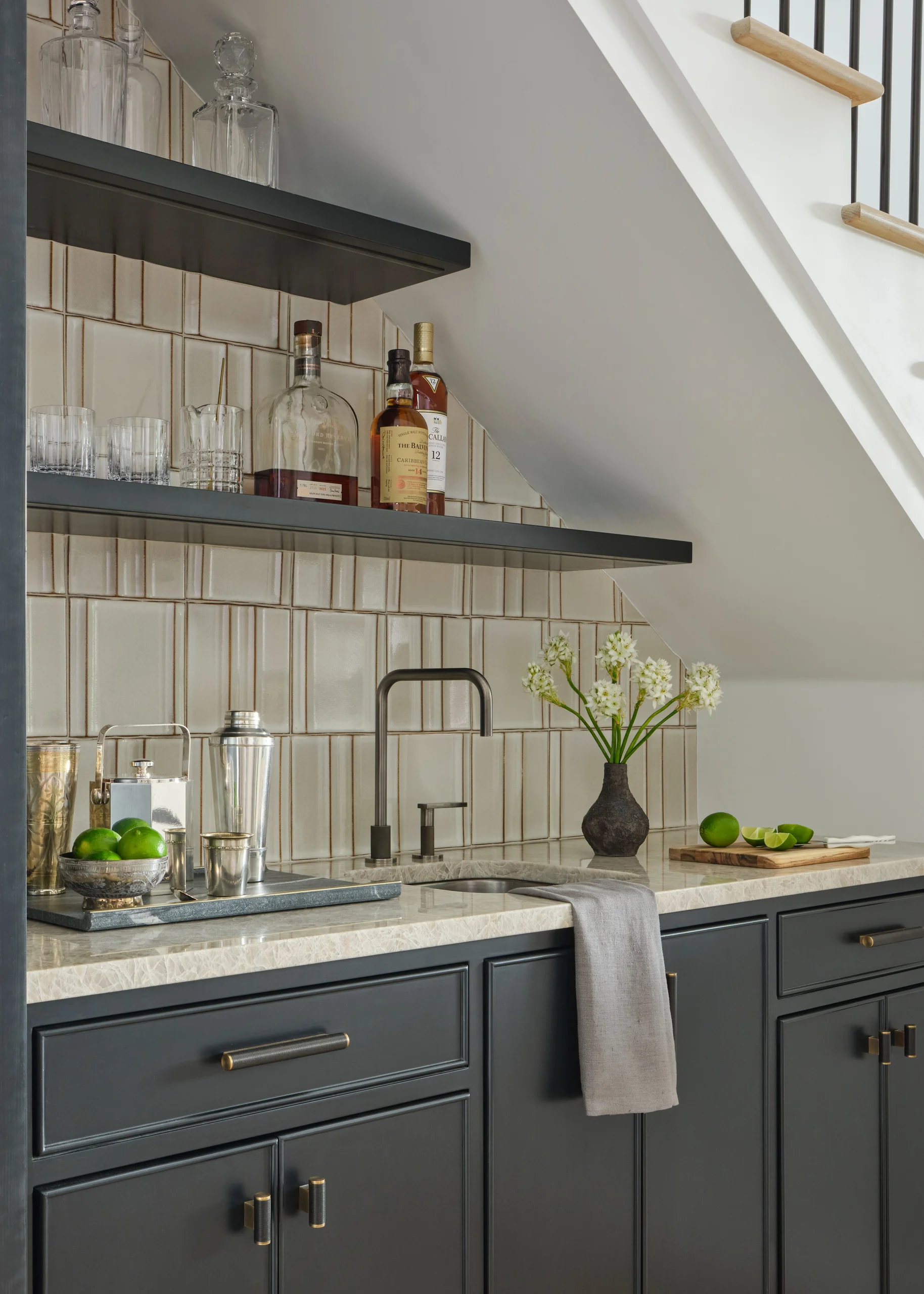 Modern kitchen counter under angled ceiling with dark cabinets, cream-colored marble countertop, a black faucet, a gray towel hanging on the sink, a vase with white flowers, a cutting board with sliced limes, two dark shelves holding glassware and liquor bottles, and a tray with green limes and metal drink accessories.