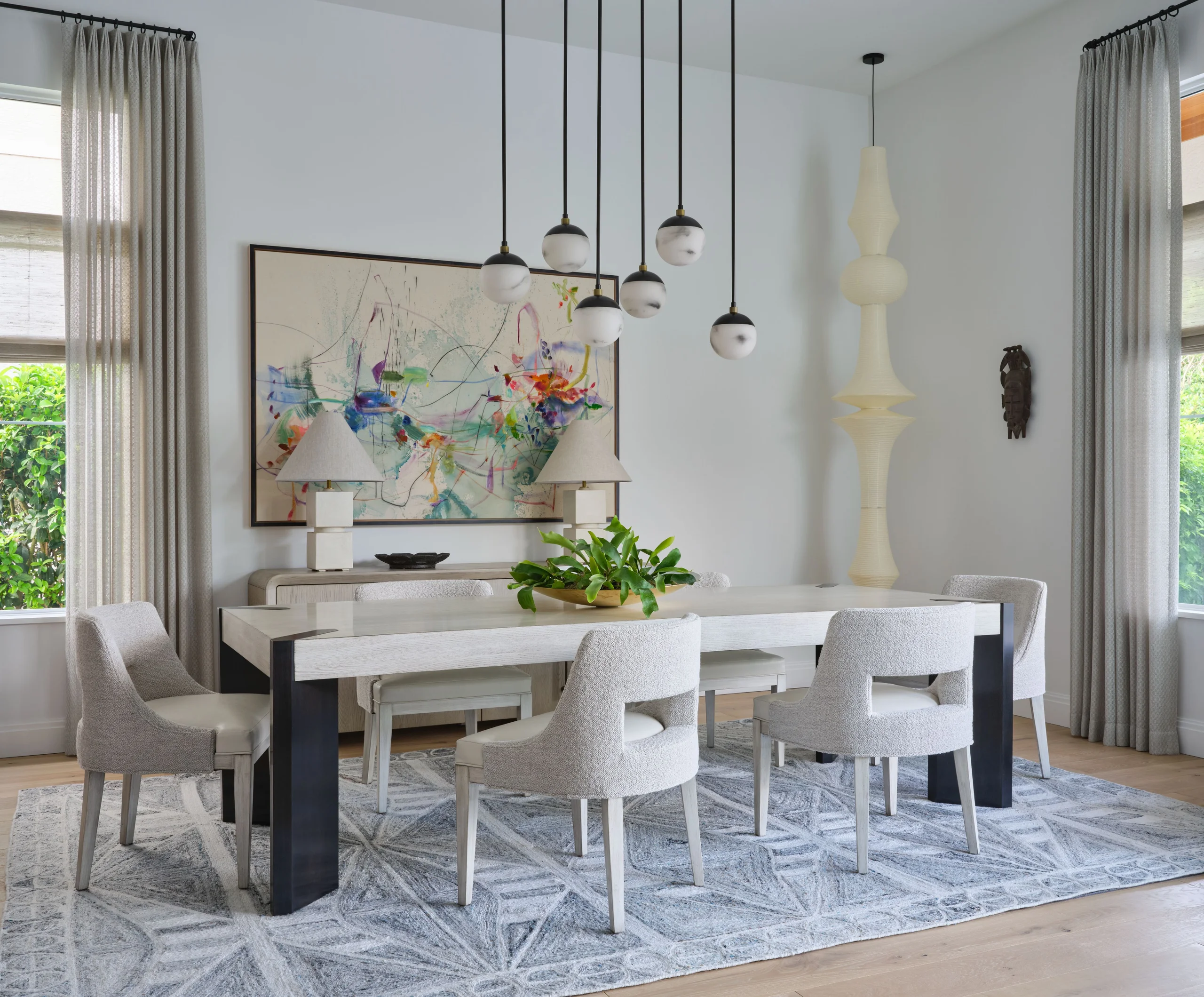 Modern dining room with a light wood table featuring black legs surrounded by six light gray upholstered chairs, a geometric patterned gray area rug, a large abstract painting on the wall, two table lamps on a sideboard, hanging pendant lights with round white-marble shades, and tall windows with sheer curtains letting in natural light.