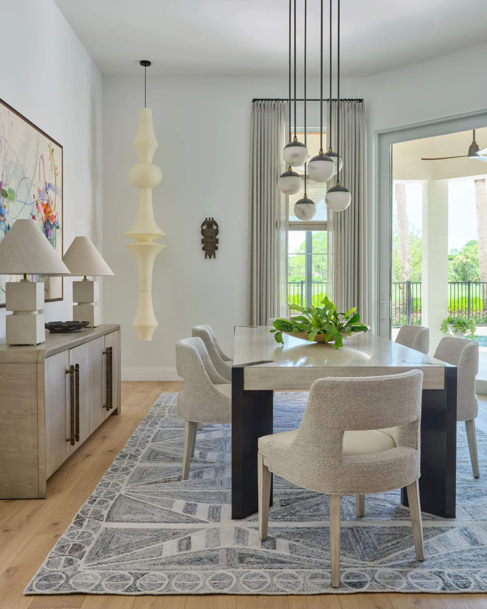 Modern dining room with a light wooden rectangular table surrounded by six beige upholstered chairs on a gray patterned rug, featuring a cluster of seven globe pendant lights hanging above the table, a sideboard with two lamps and colorful abstract art on the wall, a tall sculptural hanging lamp, and a window and sliding glass door showing greenery outside.