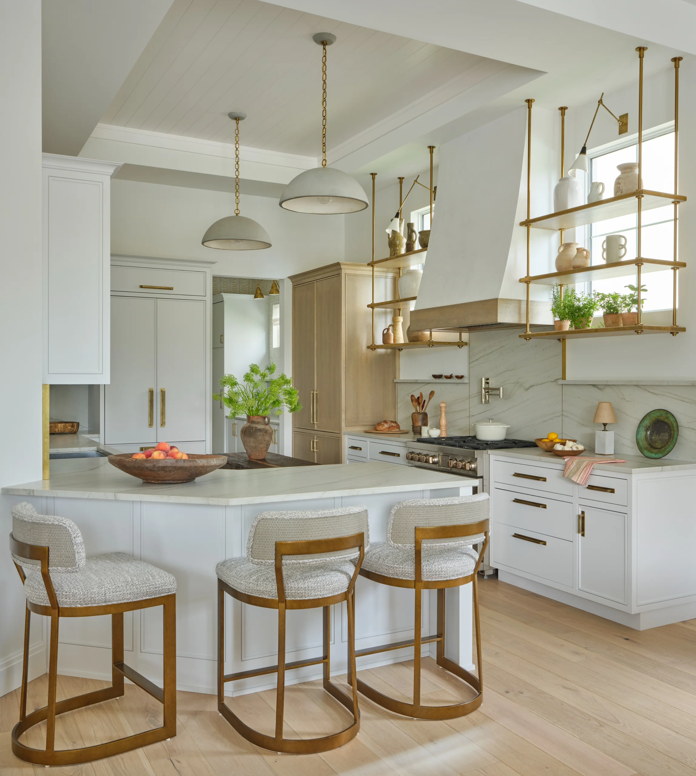 Bright modern kitchen with white cabinetry, light wood flooring, and a marble countertop island with three cushioned bar stools with brass frames, pendant lights, open brass shelving with pottery and plants, and a stove with a white hood.