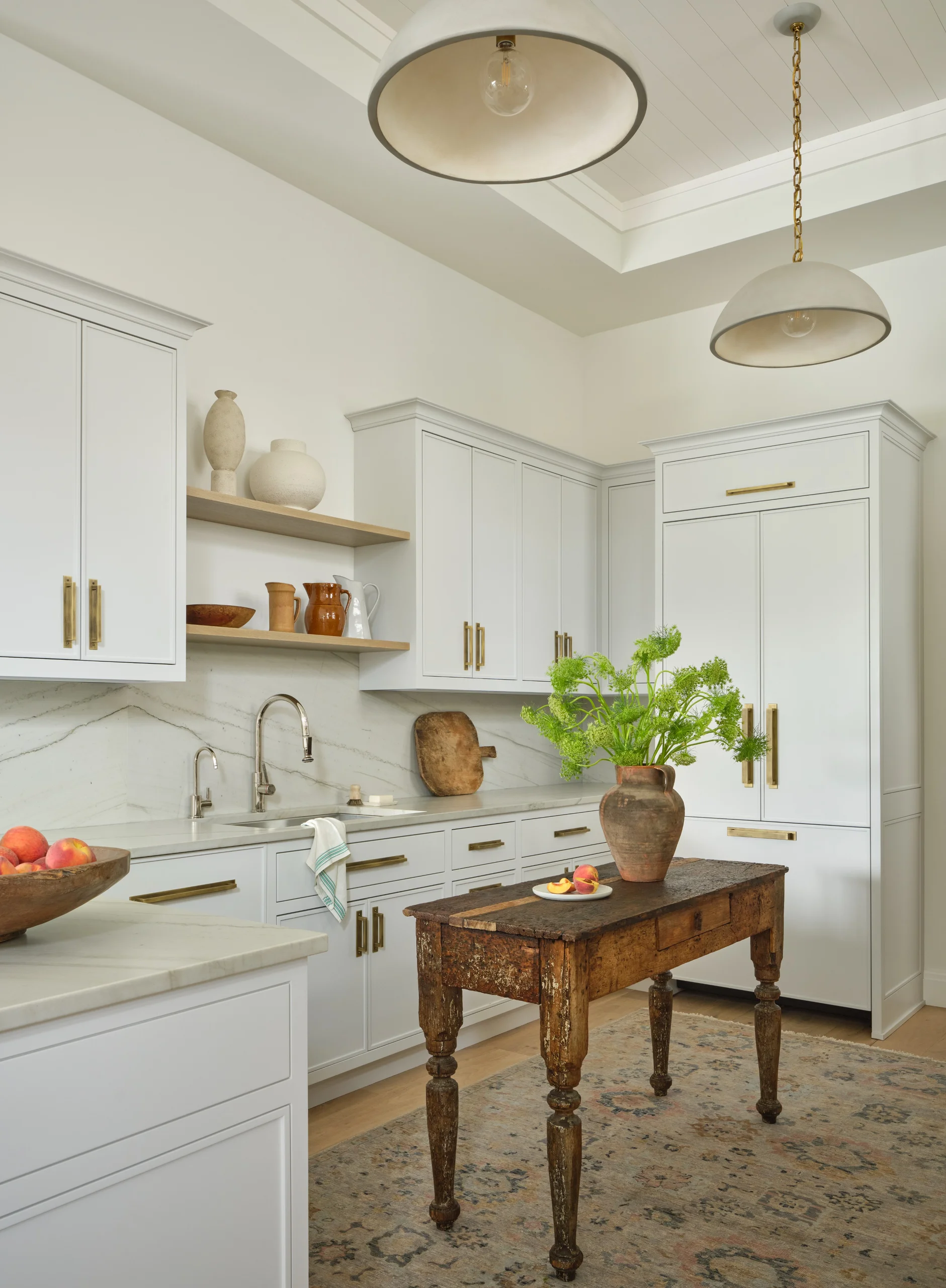Bright kitchen with white cabinetry and gold handles, marble countertops and backsplash, two pendant lights, a rustic wooden table with a vase of green flowers, and a wooden bowl of peaches on the island.