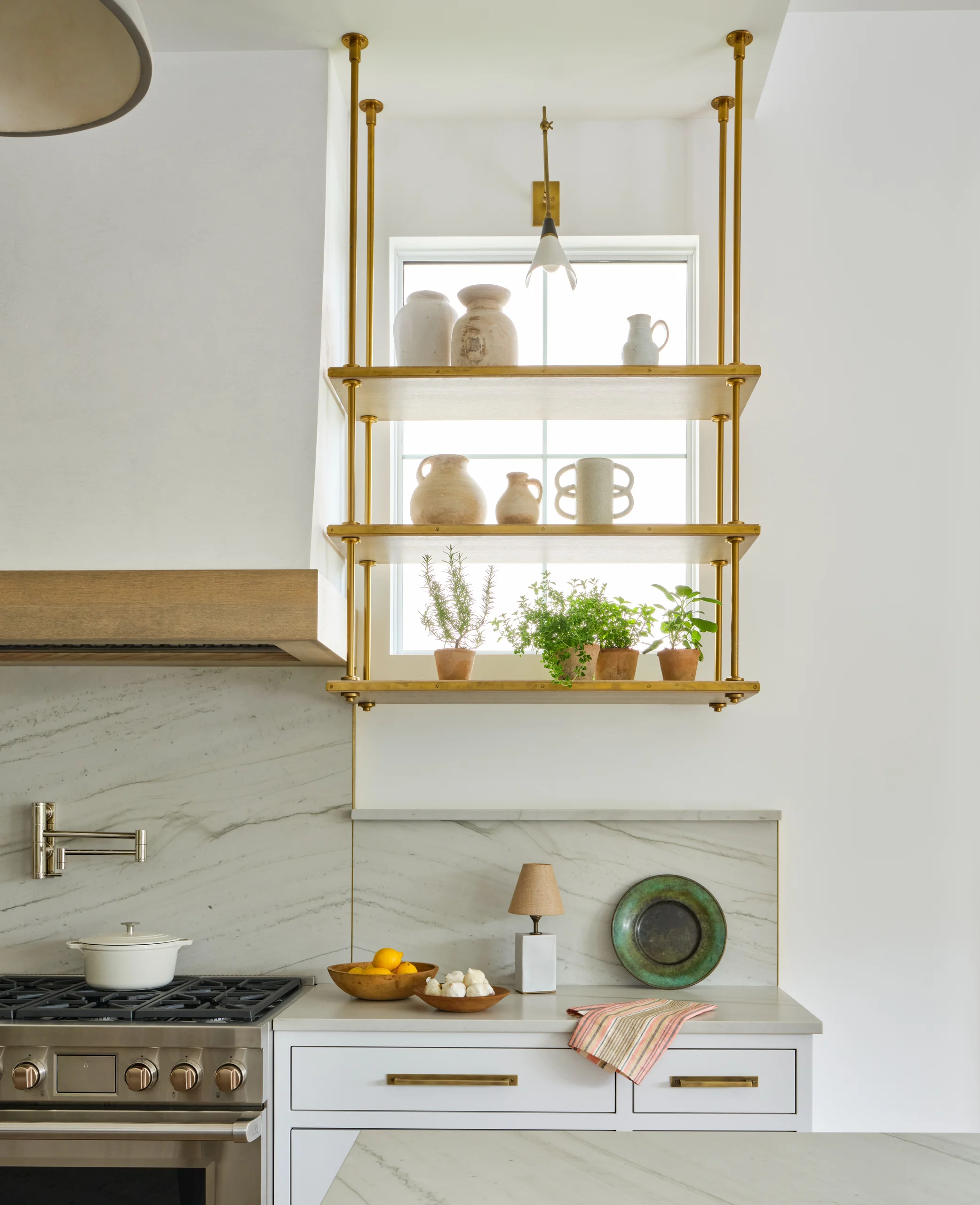 Modern kitchen corner with white cabinetry, a stainless steel stove with a white pot, marble backsplash and countertop, floating brass shelves holding ceramic jars and potted herbs, a lamp with a beige shade, a decorative green plate, wooden bowls with lemons and garlic, and a striped kitchen towel.