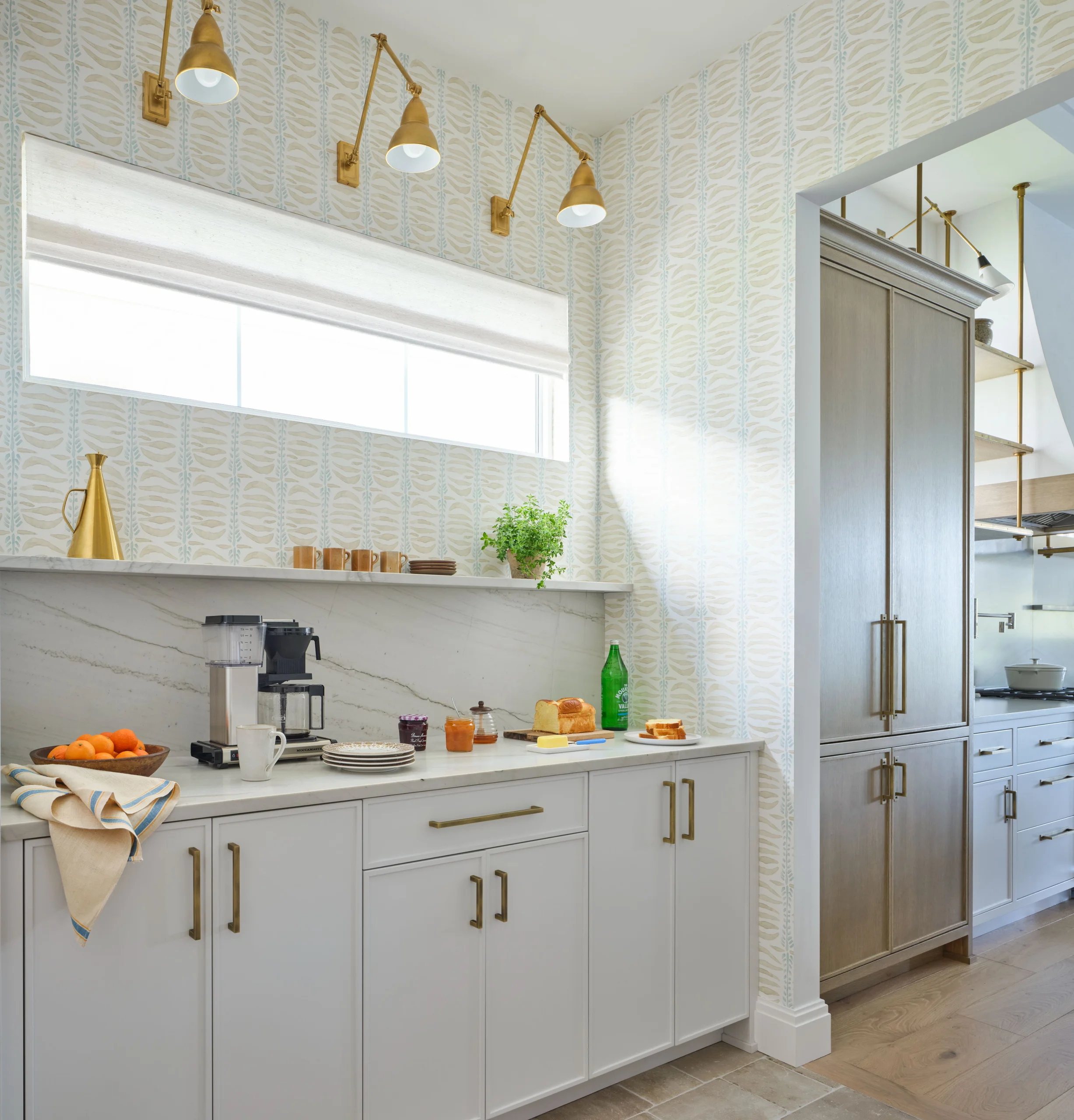 Bright kitchen corner with white cabinets, brass handles, patterned wallpaper, and a window with a white roller blind. Countertop features a coffee maker, plates, cups, a bowl of oranges, a loaf of bread with slices, jam jars, a green bottle, and a potted plant on a shelf. Brass wall-mounted lamps are above the window. Adjacent to the corner is a tall wooden cabinet with brass handles, and part of a stove and overhead brass shelving are visible.