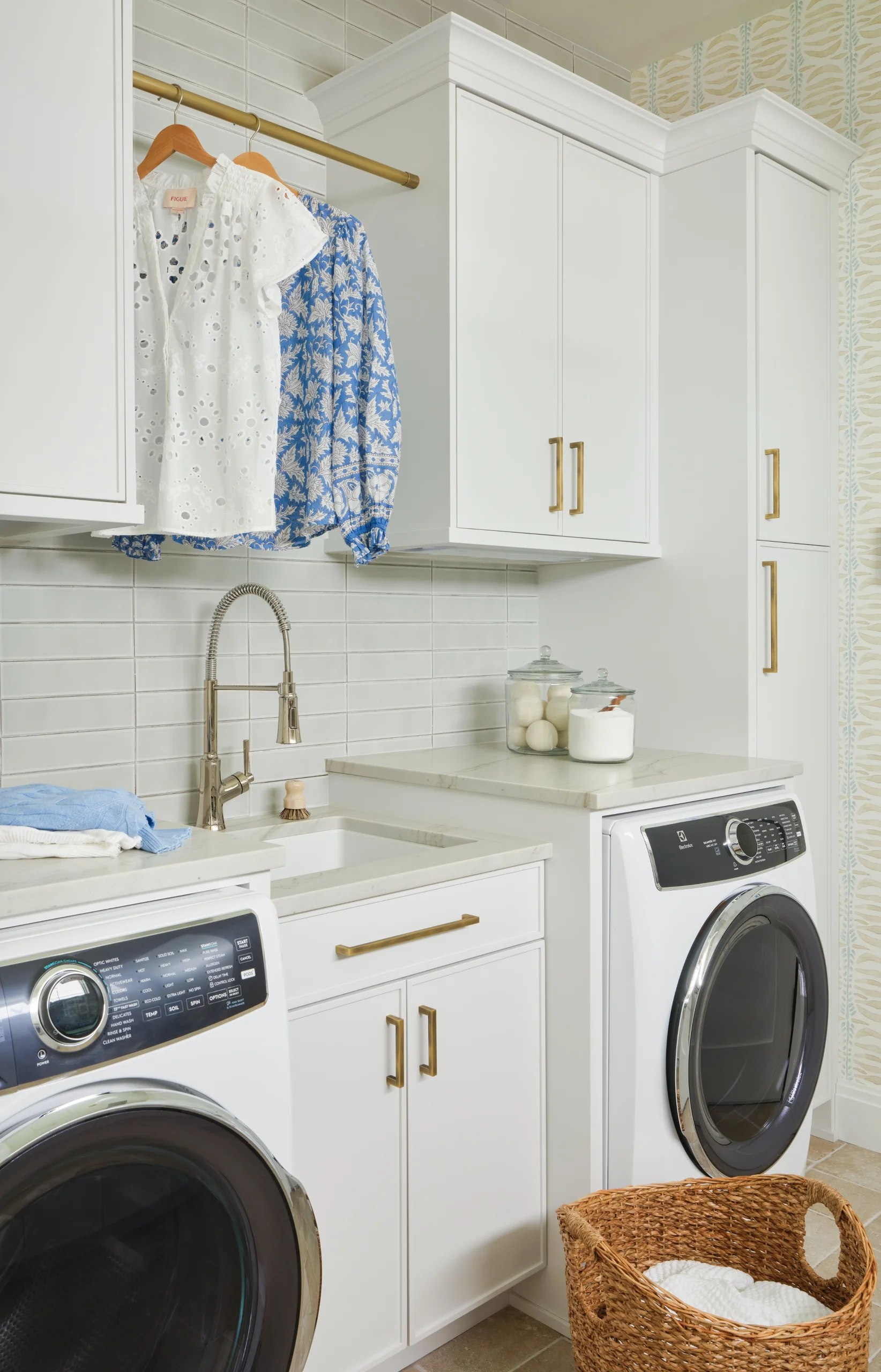 Modern laundry room with white front-loading washer and dryer under marble countertops, white cabinets with brass handles, a stainless steel faucet over a white sink, a wicker laundry basket with folded white towels, and two tops (white eyelet and blue patterned) hanging on a brass rod above the sink, against a tiled backsplash and patterned wallpaper.