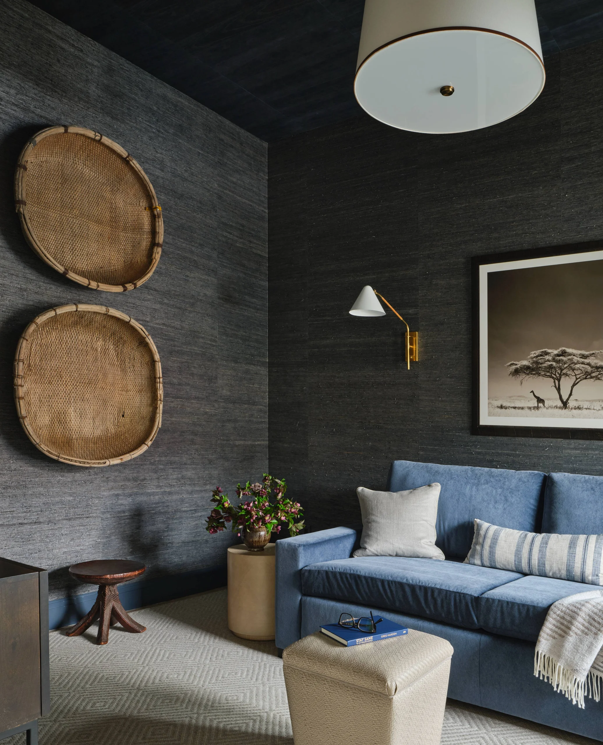 Cozy living room corner with a blue sofa adorned with two pillows and a striped throw, a beige ottoman with a book and glasses on top, two woven baskets mounted on dark textured walls, a small wooden stool, a round beige side table with a vase of flowers, a framed black and white picture of a giraffe and tree, and a white ceiling lamp.