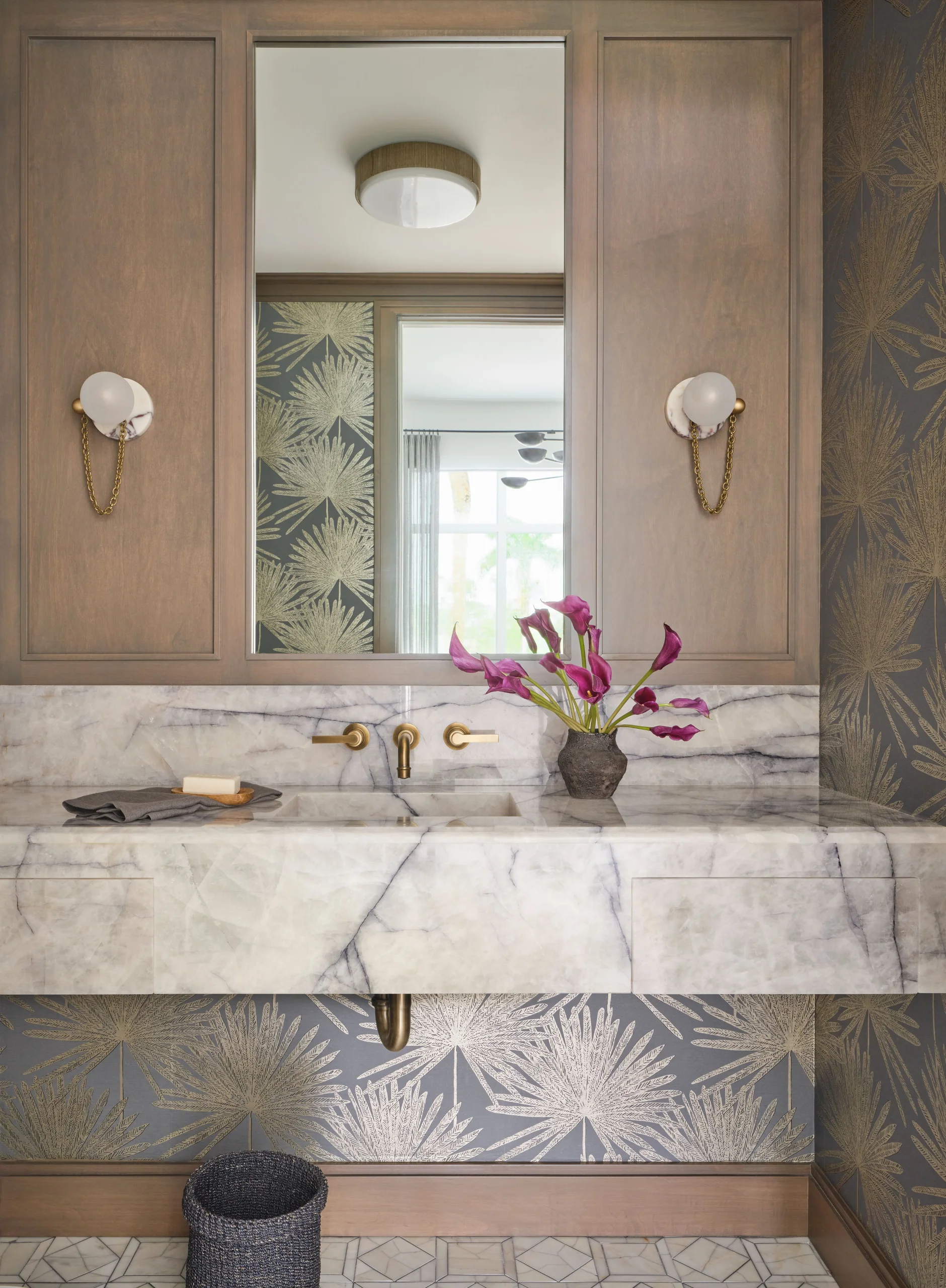 Modern bathroom vanity with white marble countertop and built-in sink, brass wall-mounted faucet, pink calla lilies in a dark vase, and a large rectangular mirror reflecting a ceiling light and window; floral patterned wallpaper in gray and gold tones, wooden panels with white globe sconces, and a small dark woven basket on patterned tile floor.